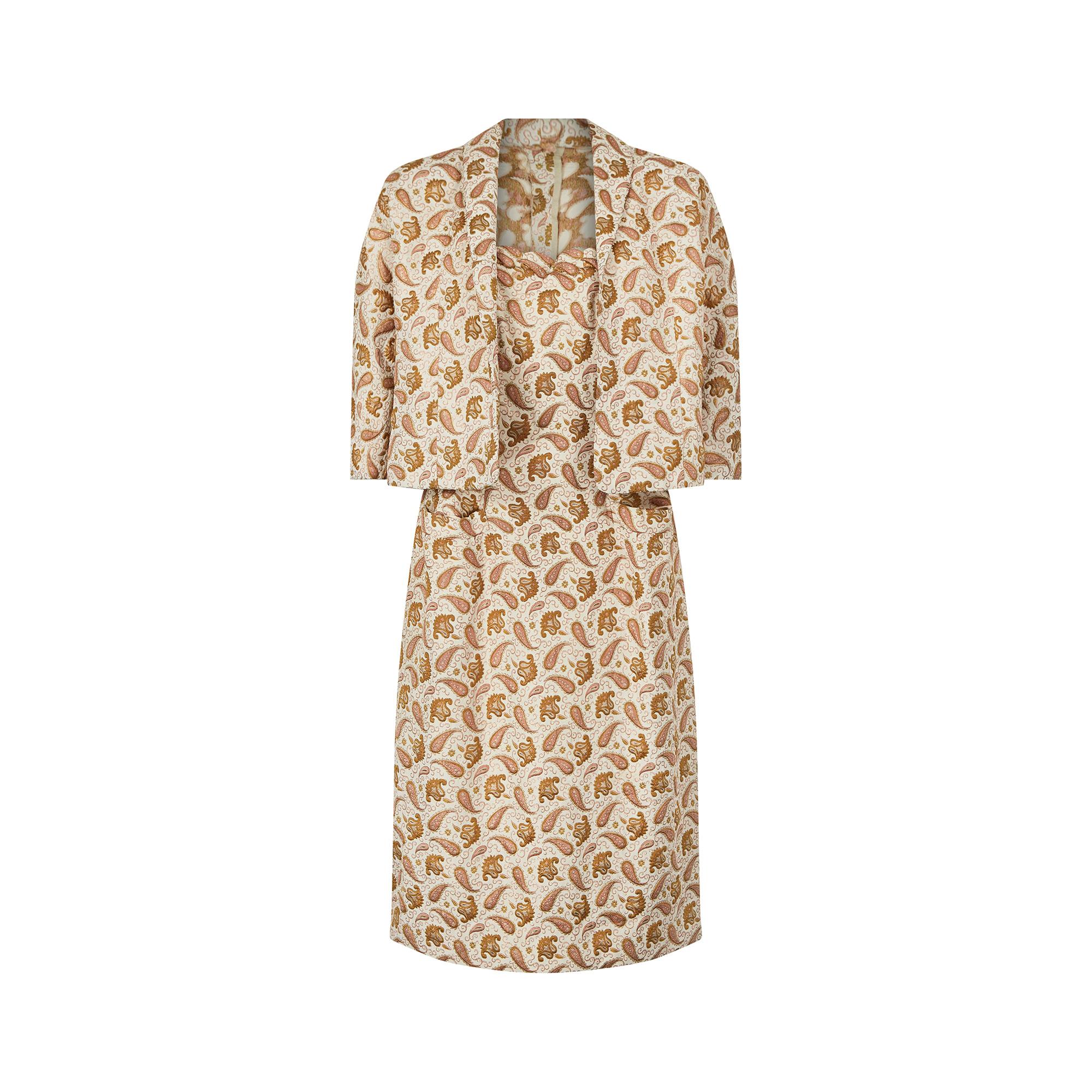 This 1950s silk brocade two piece couture dress set has a stunning paisley pattern in pink and gold threads. The dress features a scalloped edge trim to the top of the bodice and three sets of darts leading down from the bust to nip in the waist,
