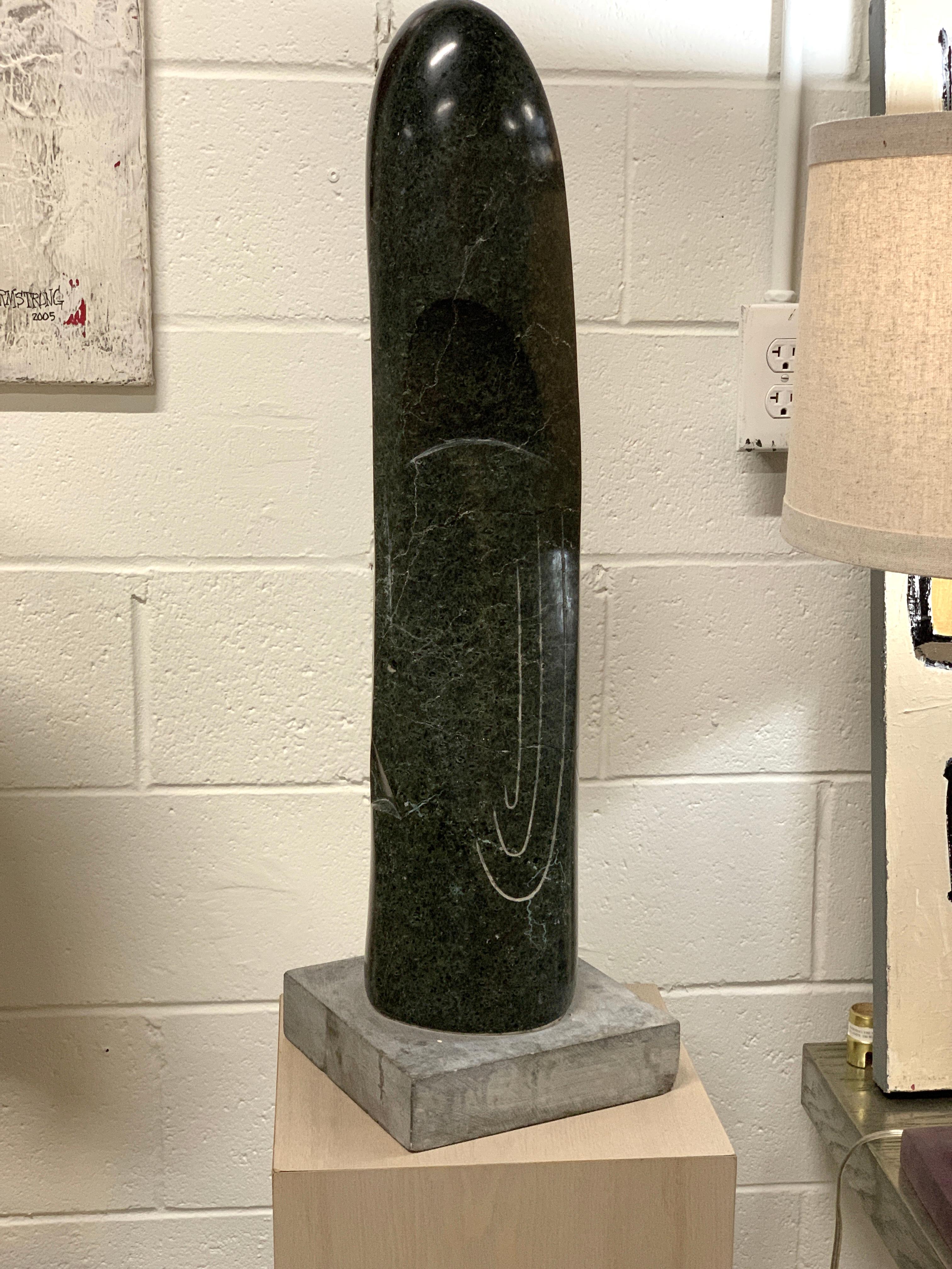 A large dark green marble dated 1957 by the Canadian artist Stanley Lewis. It is inscribed on the base Lewis 57. Depicts a face carved into the sides. The piece is quite large and heavy. There is a fracture in the middle of the sculpture which
