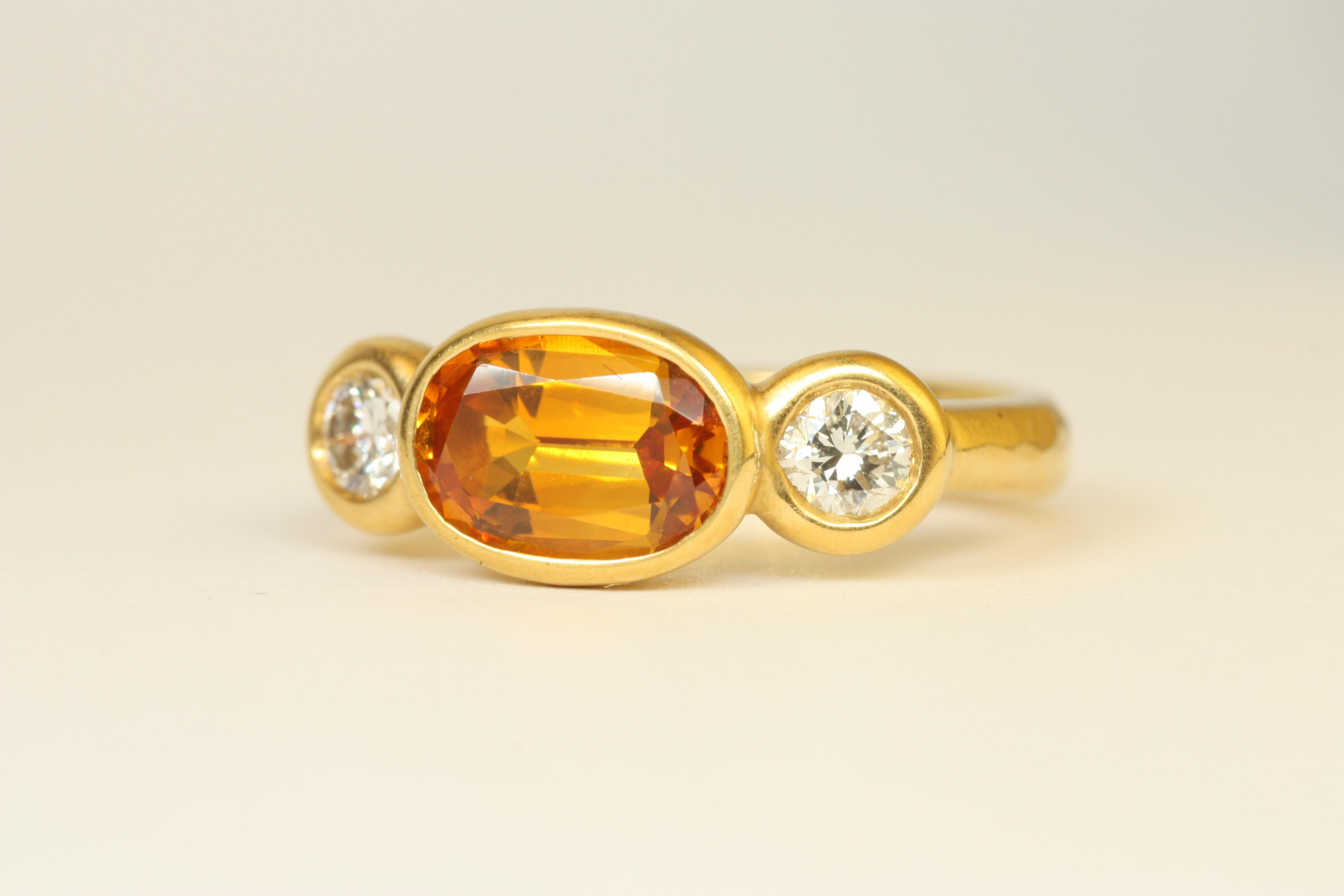 This vibrant 3 stone ring is centered with an oval yellow-brown-orange sapphire, accented by two bright round diamonds, all set in rich 22k yellow gold. The hammered bezel setting brings out the warm tones of the stones, creating a piece that’s both