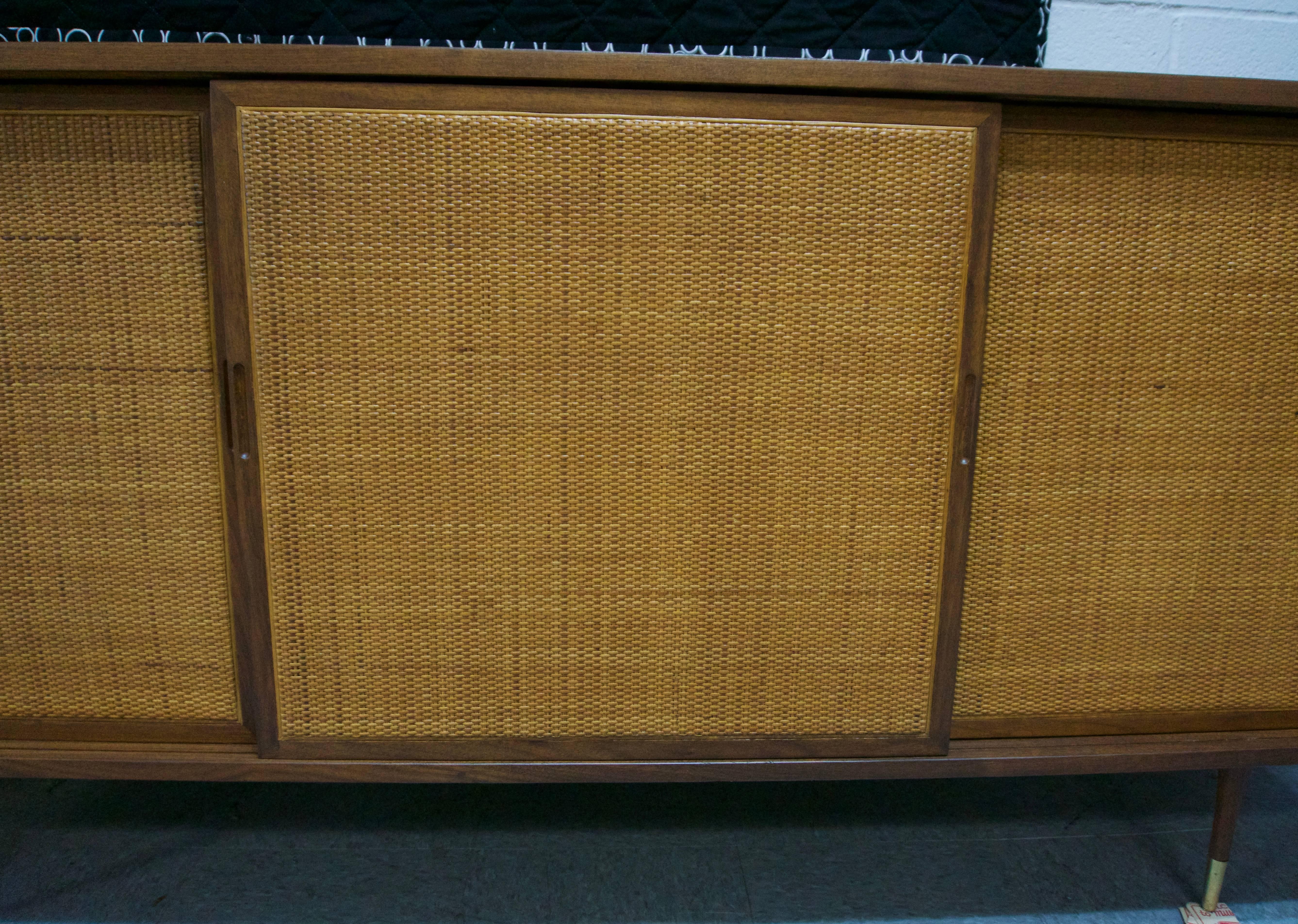 Restored walnut credenza with sliding woven cane doors. The interior is black with four natural drawers and two shelves. Brass tips on the feet.