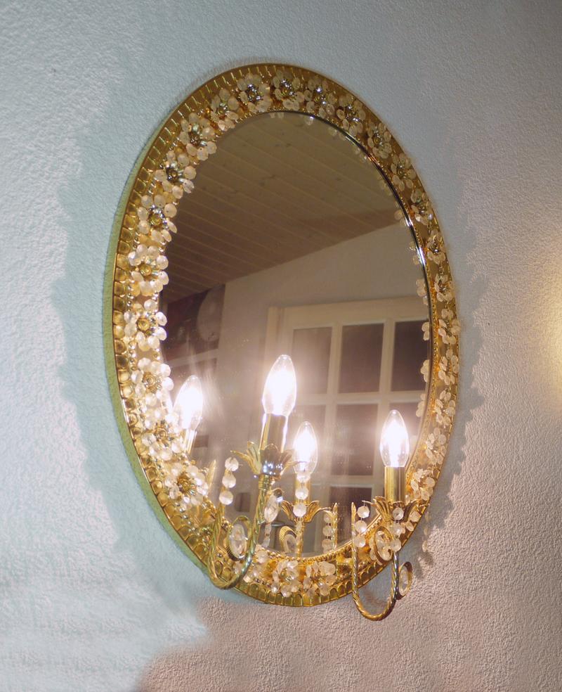 Elegant oval wall-mounted illuminated mirror with two arms and crystals flowers on a gilded brass frame. 

Manufactured by Palwa, Palme & Walter, Germany in the 1960s. 

Style: Vintage, Hollywood Regency, Mid-Century Modern. 
Colours: gold,