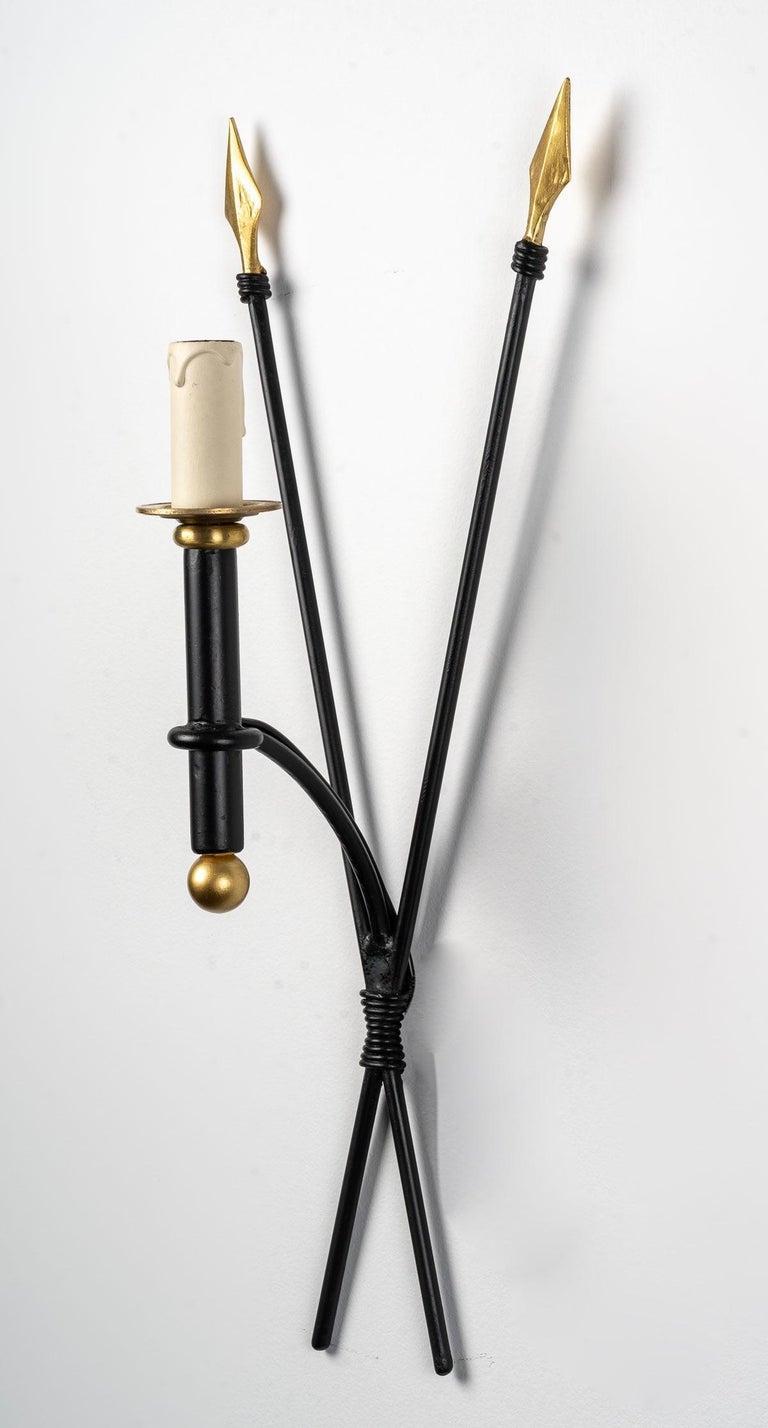 Composed of two black wrought iron arrows decorated with golden feathers, they cross on the lower part.
At the intersection of the arrows, an arm of light placed at the front of the wall lamp is formed by a black wrought iron tube adorned with a