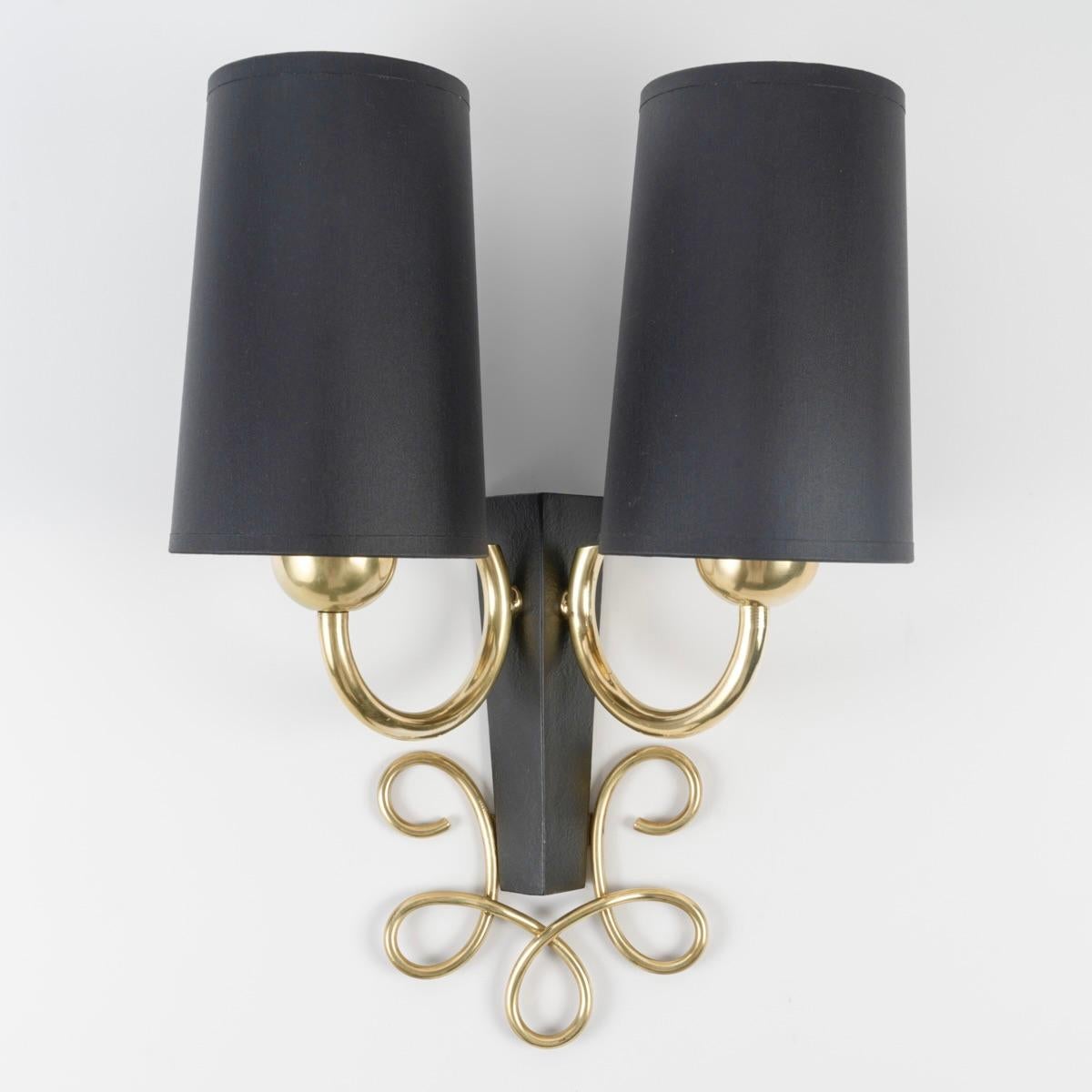 Consta de un soporte de pared trapezoidal de hierro forjado negro que sostiene dos brazos de luz de latón dorado en la parte superior, cubiertos con pantallas trapezoidales negras idénticas hechas a medida.
Justo debajo, el aplique está adornado con