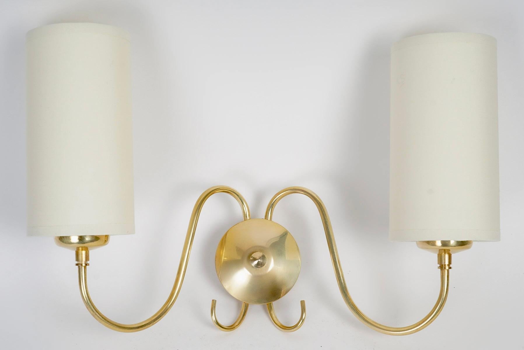 Composed of a round wall plate on which two upward-facing light arms are positioned on either side of the sconce, highlighted at the bottom by two gilded brass half-loops.
The two light arms are clad with cylindrical lampshades in off-white