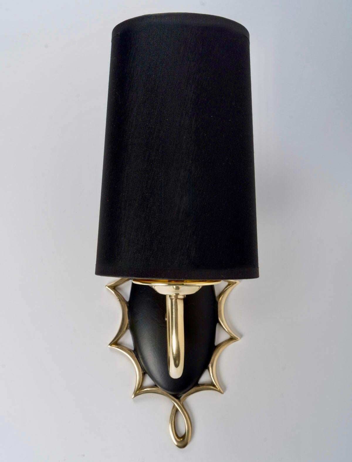 Si tratta di un'applique a forma di medaglione in bronzo annerito, ornata da un filo d'ottone che si snoda lungo il perimetro.
Al centro del medaglione, un braccio in ottone dorato è rivestito da un paralume trapezoidale, dorato all'interno e nero