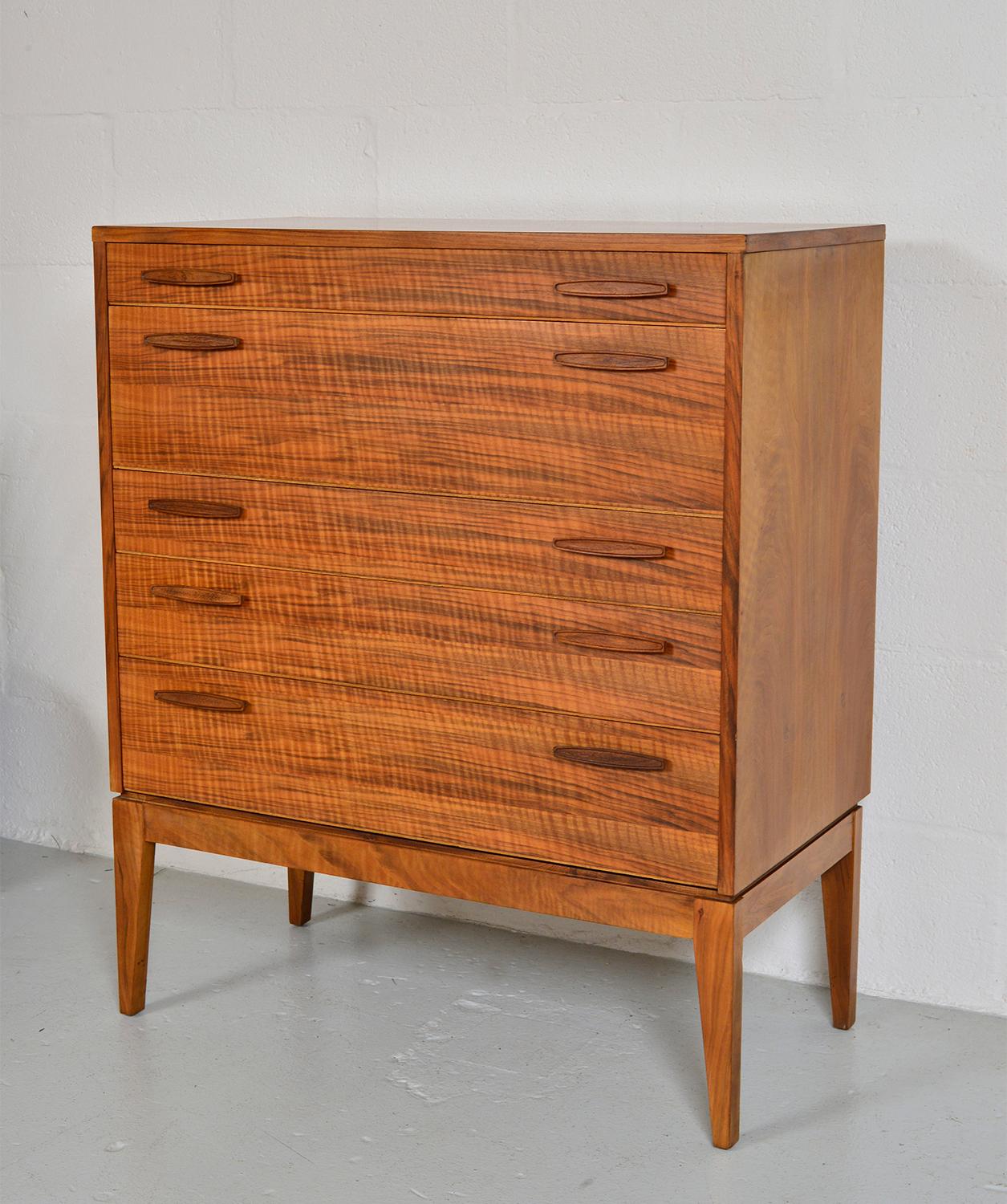 Stunning midcentury chest of drawers in well-figured walnut by renowned British furniture maker, Alfred Cox, supplier to Heals of London. The chest of drawers stands on gently tapered legs, with five dovetailed drawers, of various depths. The