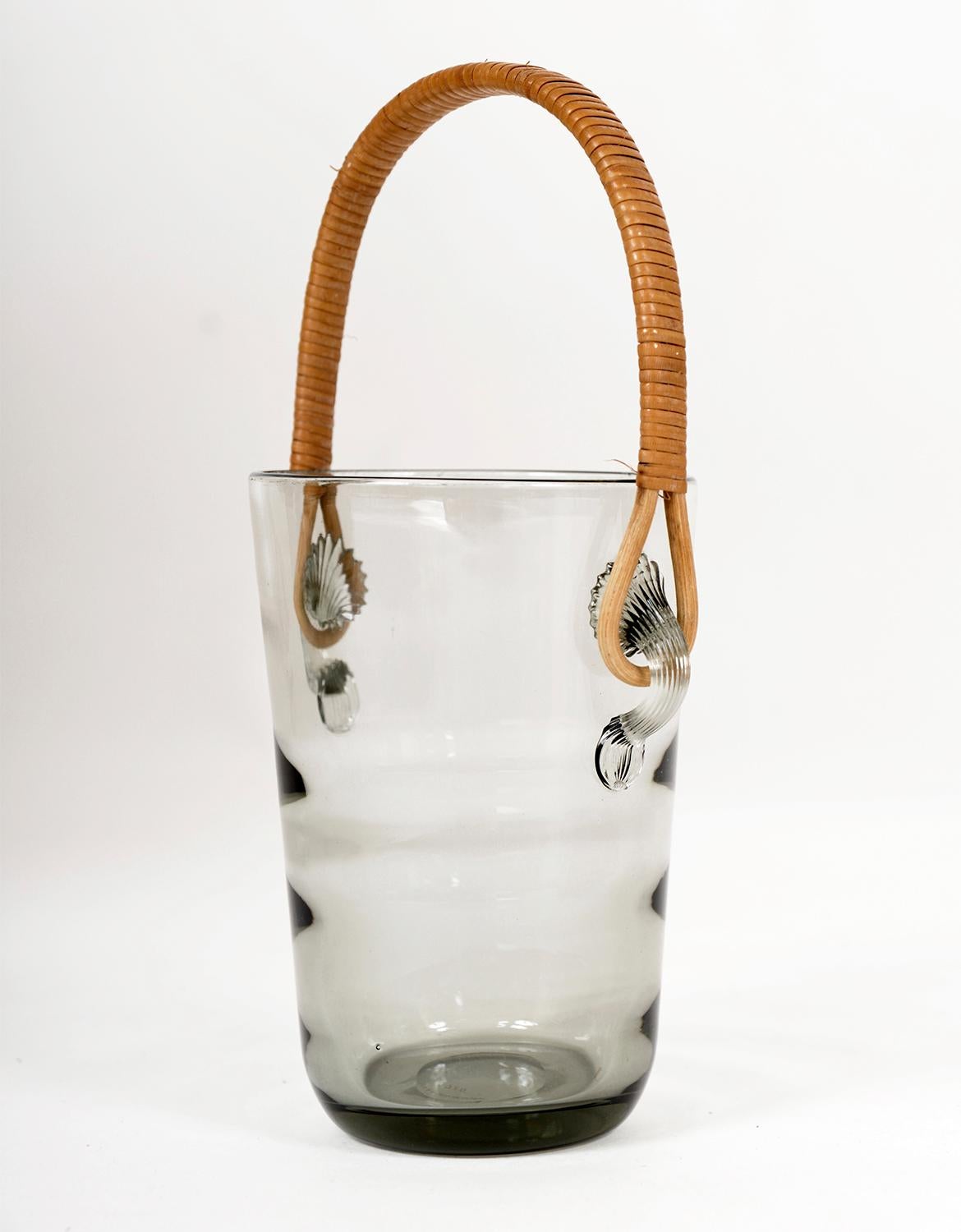 Per Lutken designed this very stylish “Akva” ice bucket with a slightly ribbed smoked glass pattern and an elegant rattan handle. 
Made by the famous Danish glassmakers Holmegaard in the late 1950s, early 60s, it carries the maker’s mark 