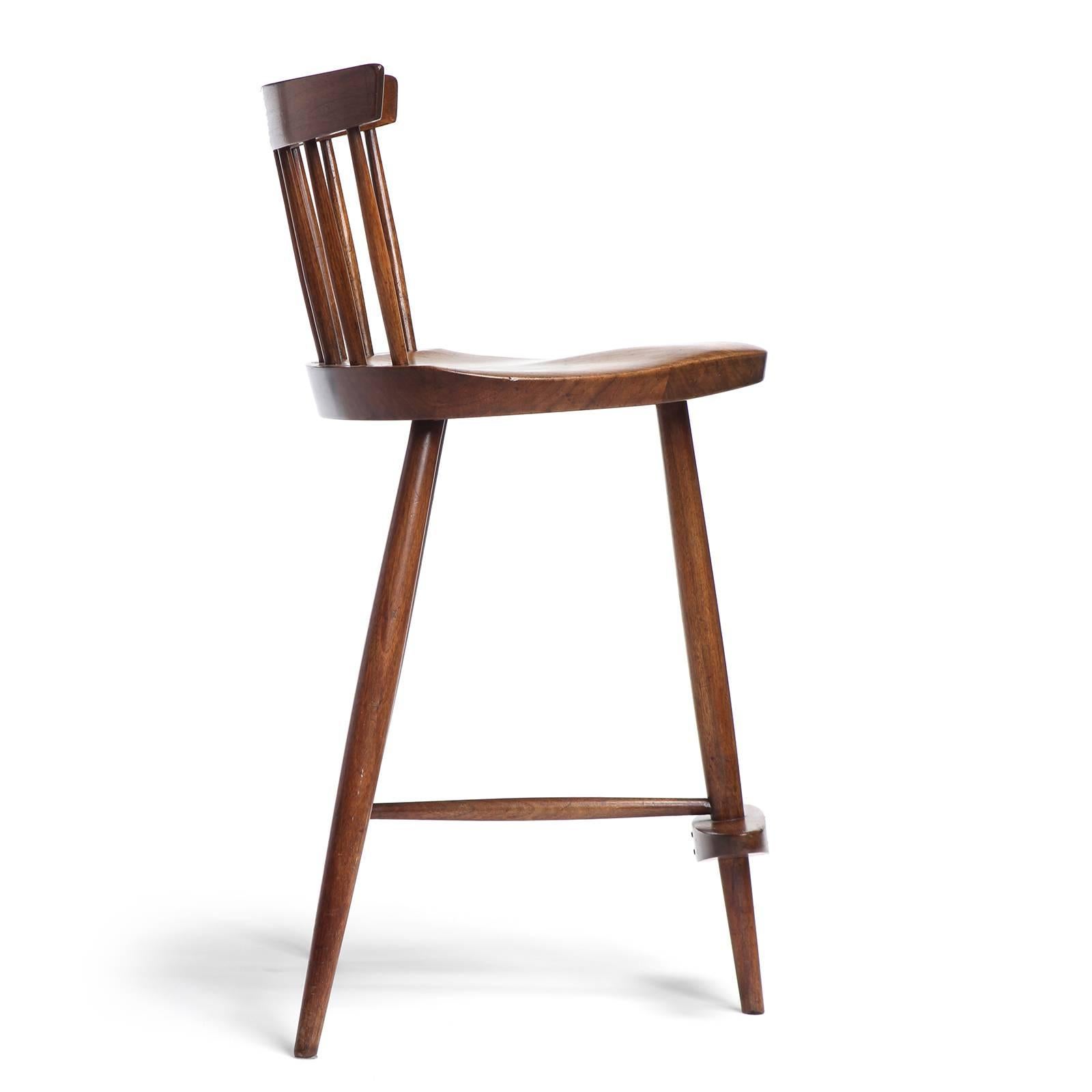 A bench-made walnut Mira stool by George Nakashima, father of the American Craft movement. The stool features hand-hewn spindles supporting a thin backrest and a shaped triangular seat on three tapered dowel legs with a central footrest.
Produced