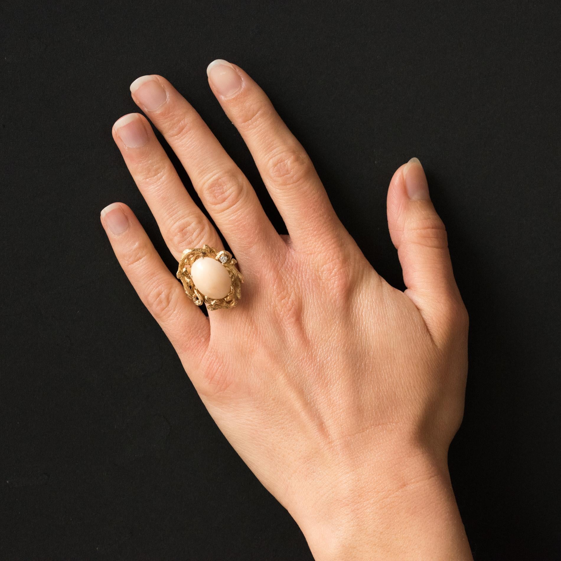 Ring in 14 karat yellow gold.
This beautiful ring with asymmetrical lines is set with claws of an angel skin coral cabochon in a gnarled decor, chiseled like a branch that frames the gem. A brilliant- cut diamond is set with claws on one