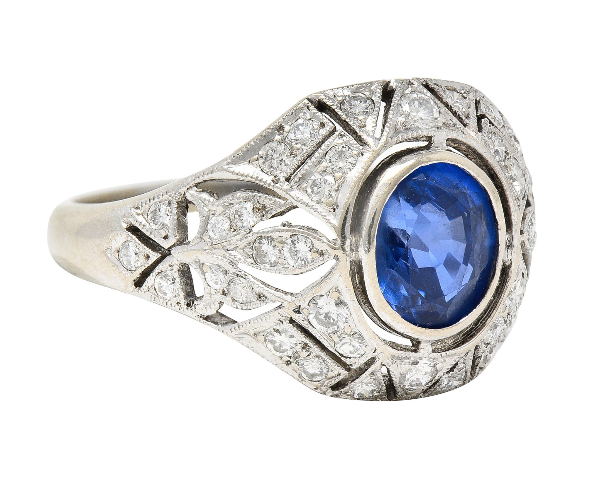 Centering an oval cut sapphire weighing approximately 1.20 carats - transparent medium blue. Bezel set with a pierced geometric surround and fleur-de-lis motif shoulders. Featuring round brilliant cut diamonds bead set throughout. Weighing