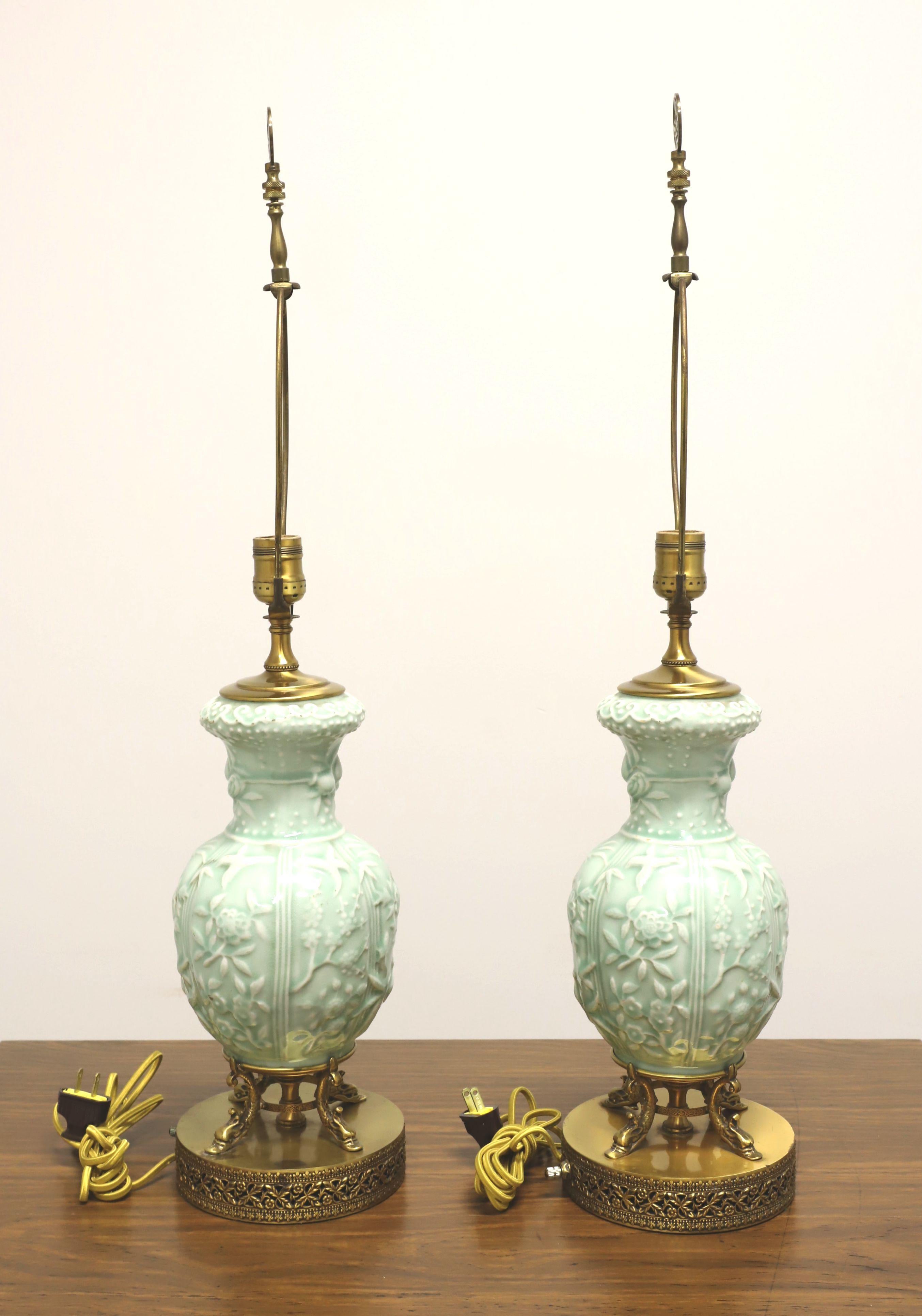 A pair of Asian style table lamps, unbranded. Made of porcelain in an urn style, celadon glaze of blue/green/white color, raised foliate Chinoiserie pattern, and on four decorative brass dolphins atop a round filigree base. Has removable metal harps