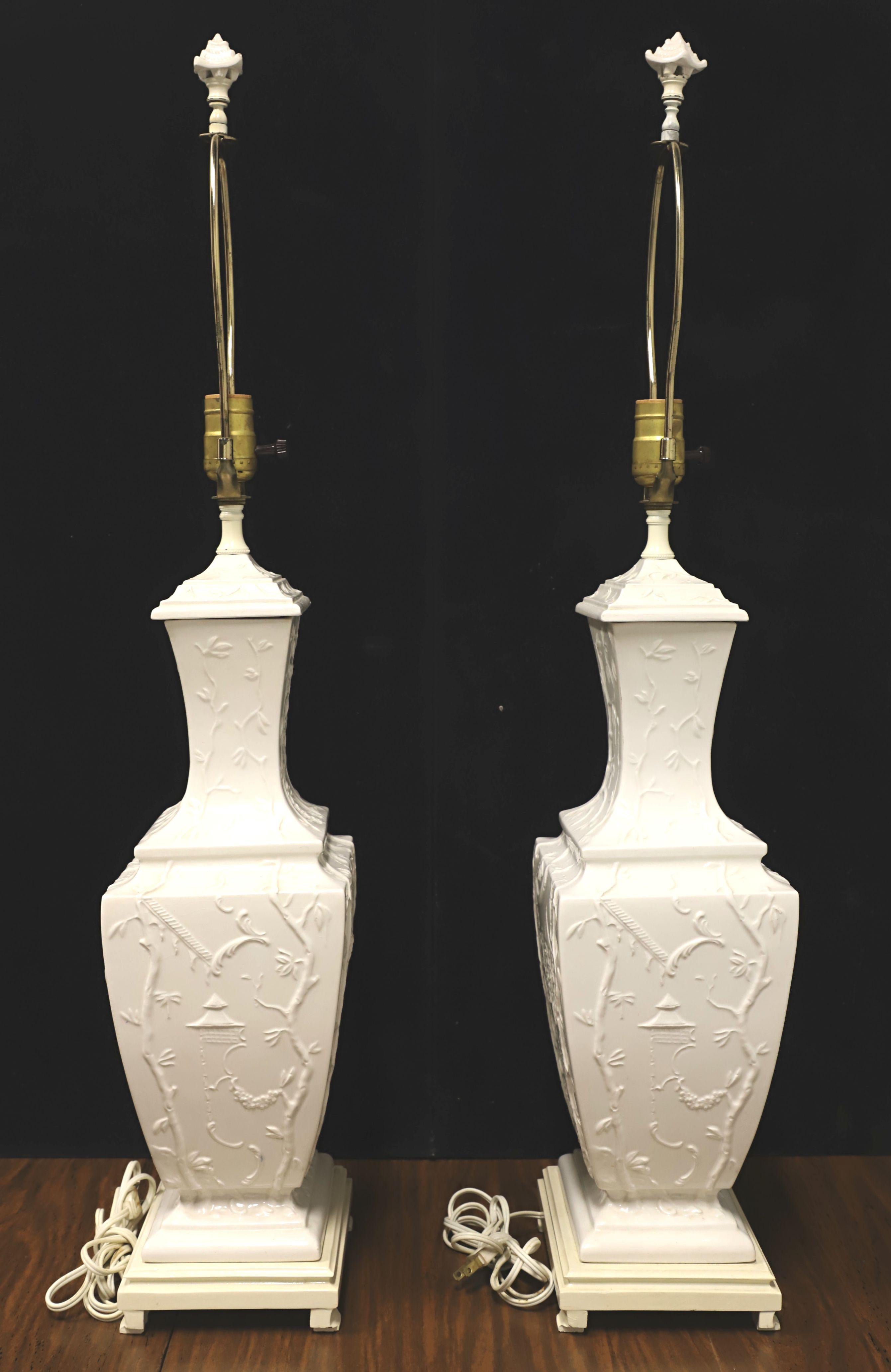 A pair of Asian Chinoiserie style table lamps, unbranded. Body is made of porcelain in an urn style, creamy white in color, raised floral Chinoiserie scene, and on a painted ceramic base. Has removable metal harps and decorative porcelain pagoda