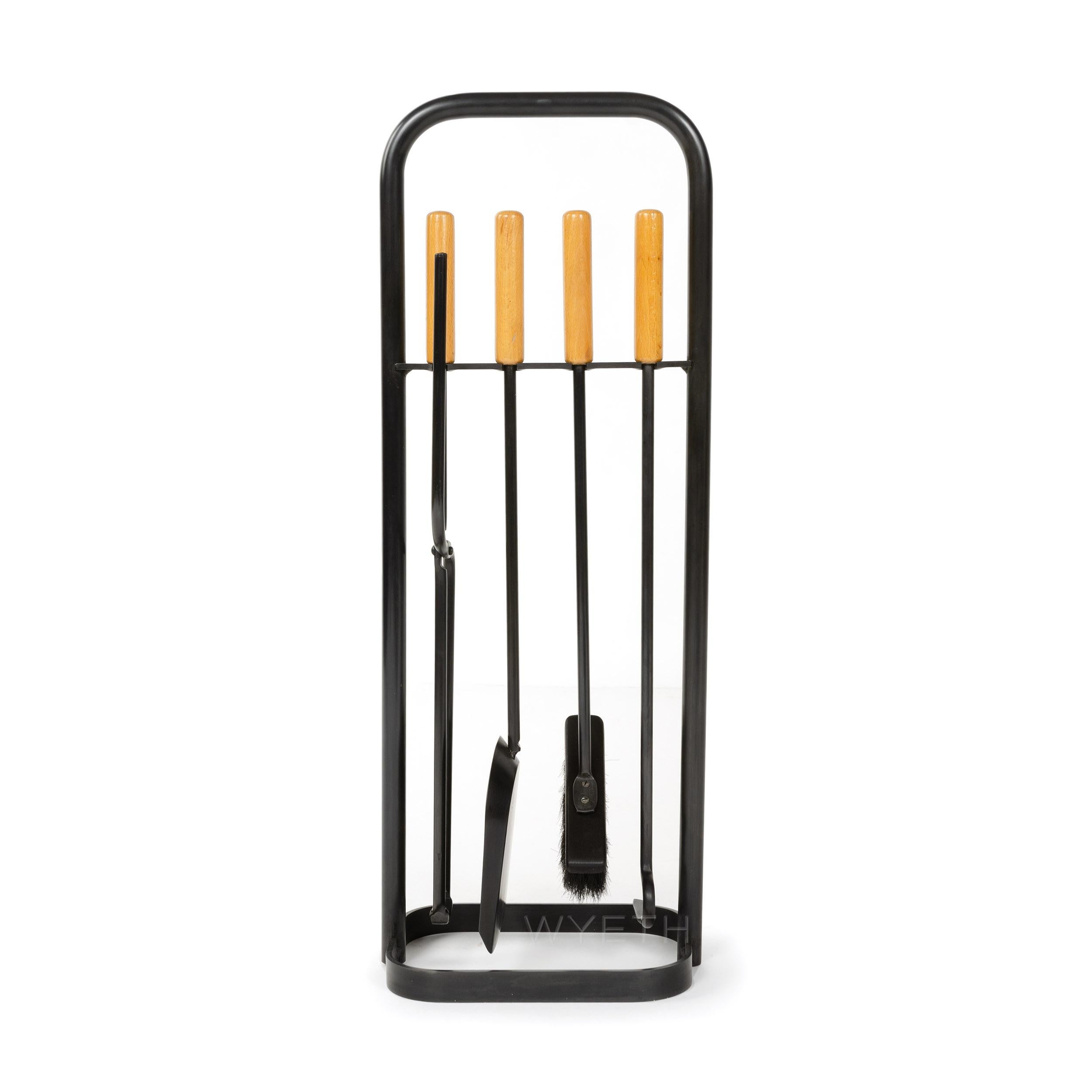 A set of handmade iron fire tools with a patinated steel finish and natural wood handles, on an inline stand with a rectangular base.