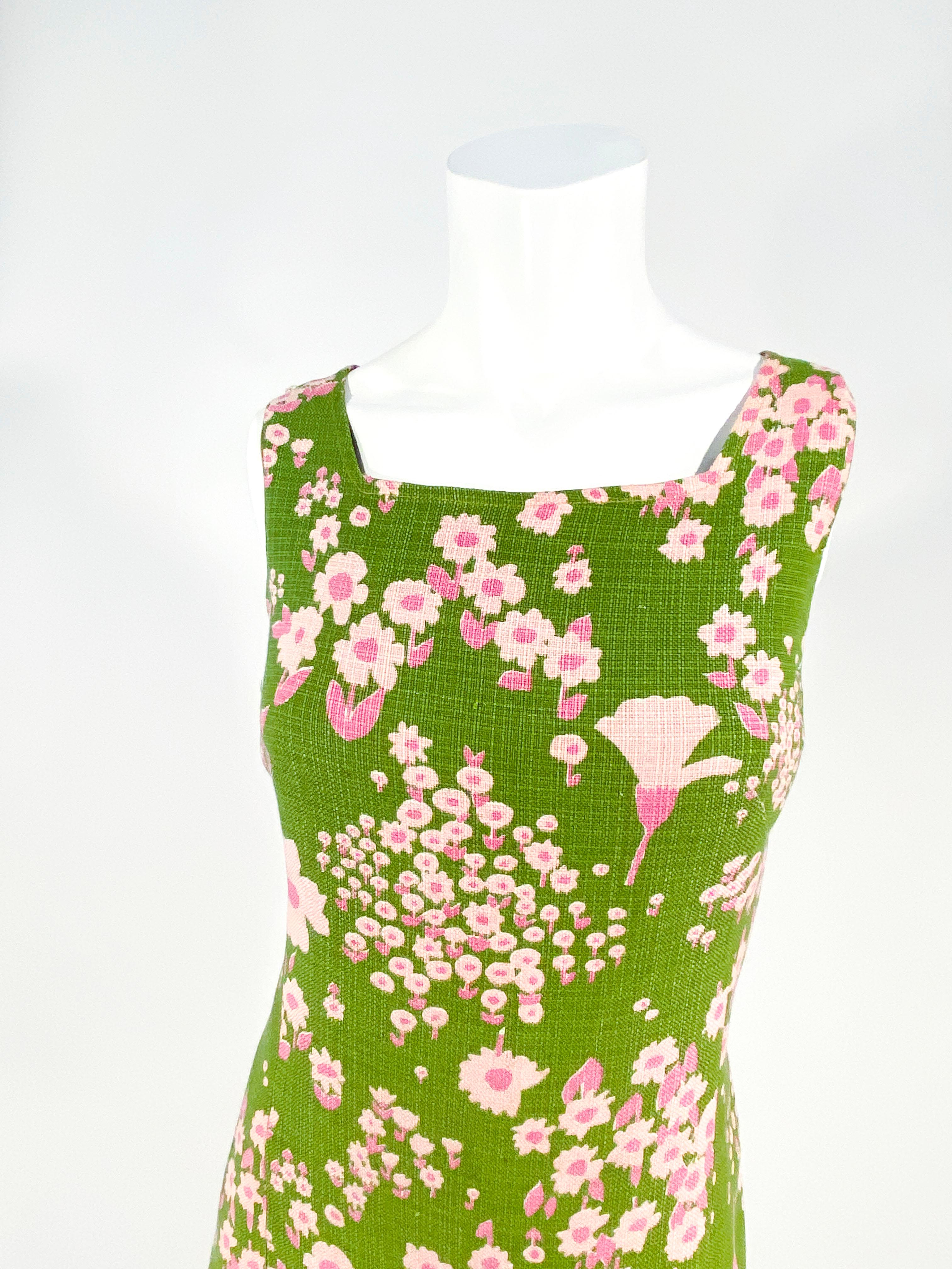 1960s avocado green cotton-linen blend day dress featuring a floral print in two shades of pink. The skirt of the dress is full at the drop-waist. This dress is fully lined, sleeveless, and has a metal back zipper. 