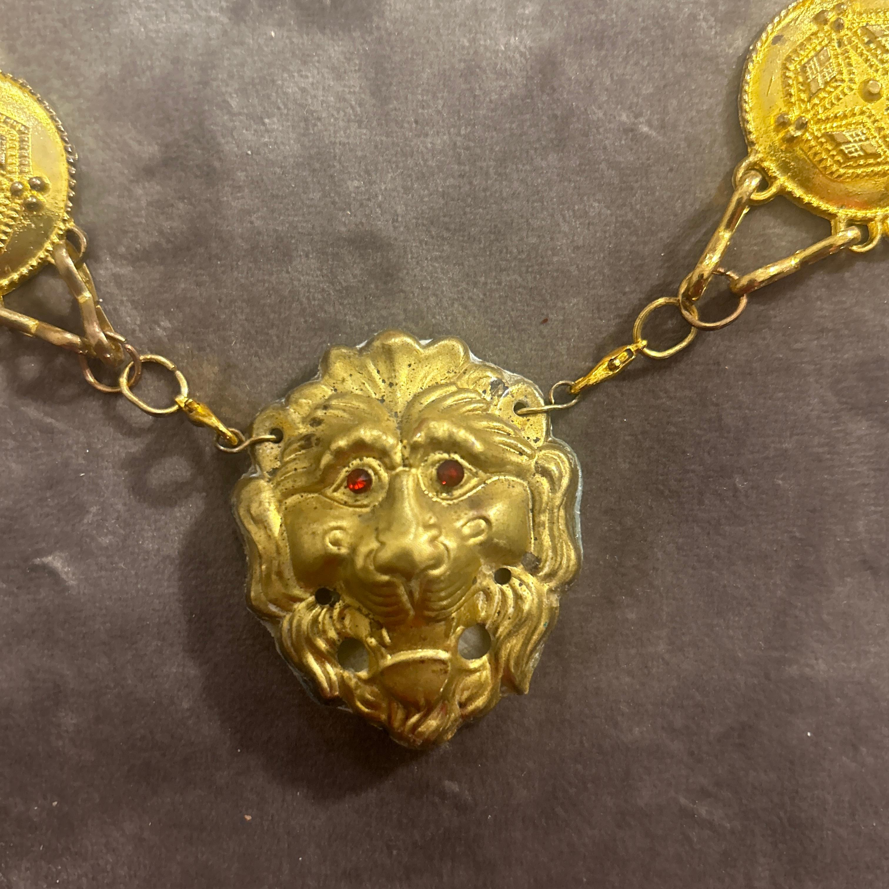 This necklace from the 1960s is a piece of costume jewelry with a bold, gold-tone design. It features a chain of round medallions with intricate details connected by smaller links. At the center of the necklace is a lion's head, which serves as a