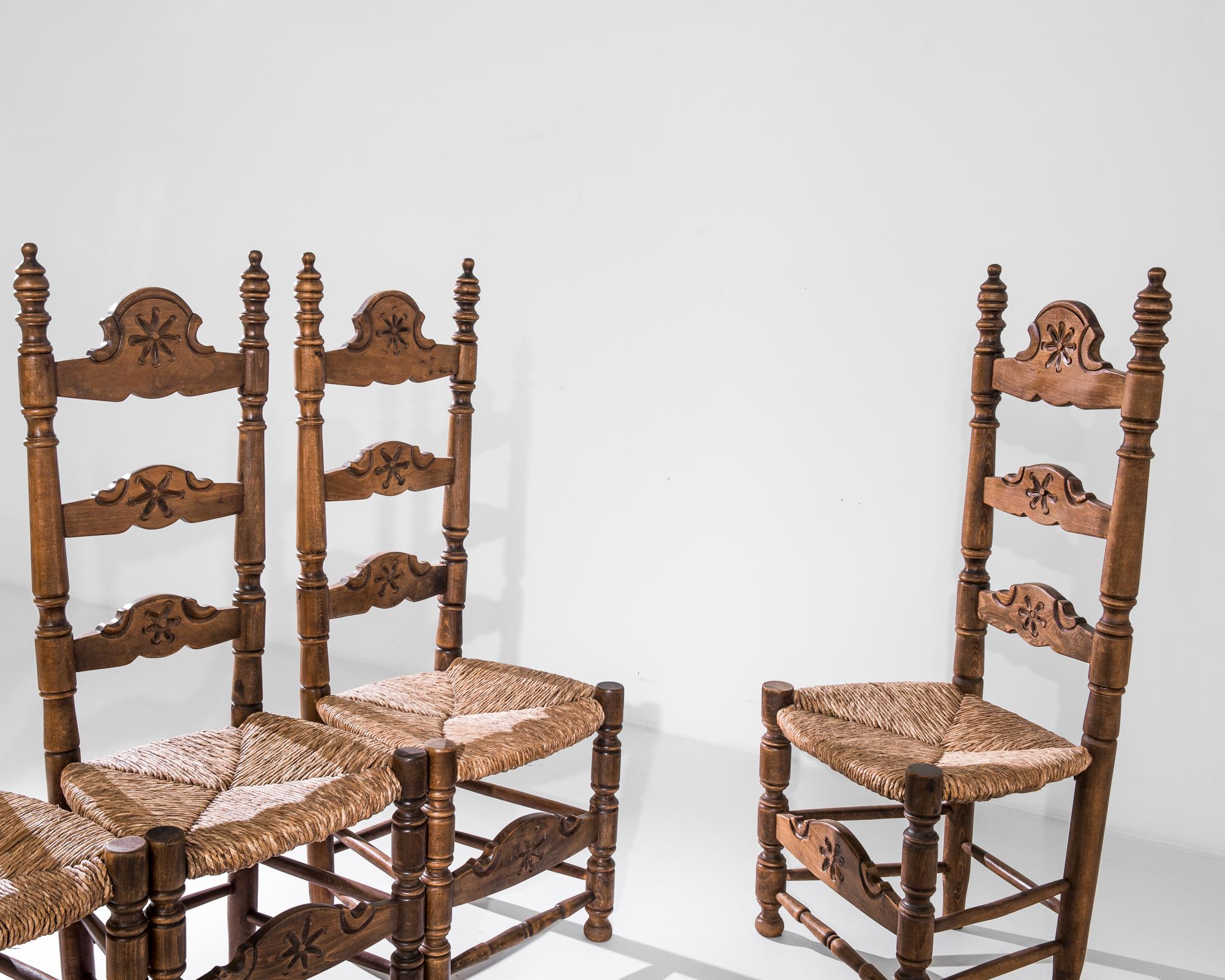 Crafted in Belgium circa 1960, this set of wooden dining chairs imparts the air of regal dignity. The chairs feature high backrests with finials, light-toned wicker seats and skilfully carved baluster legs. The adorable flower motif echoed on