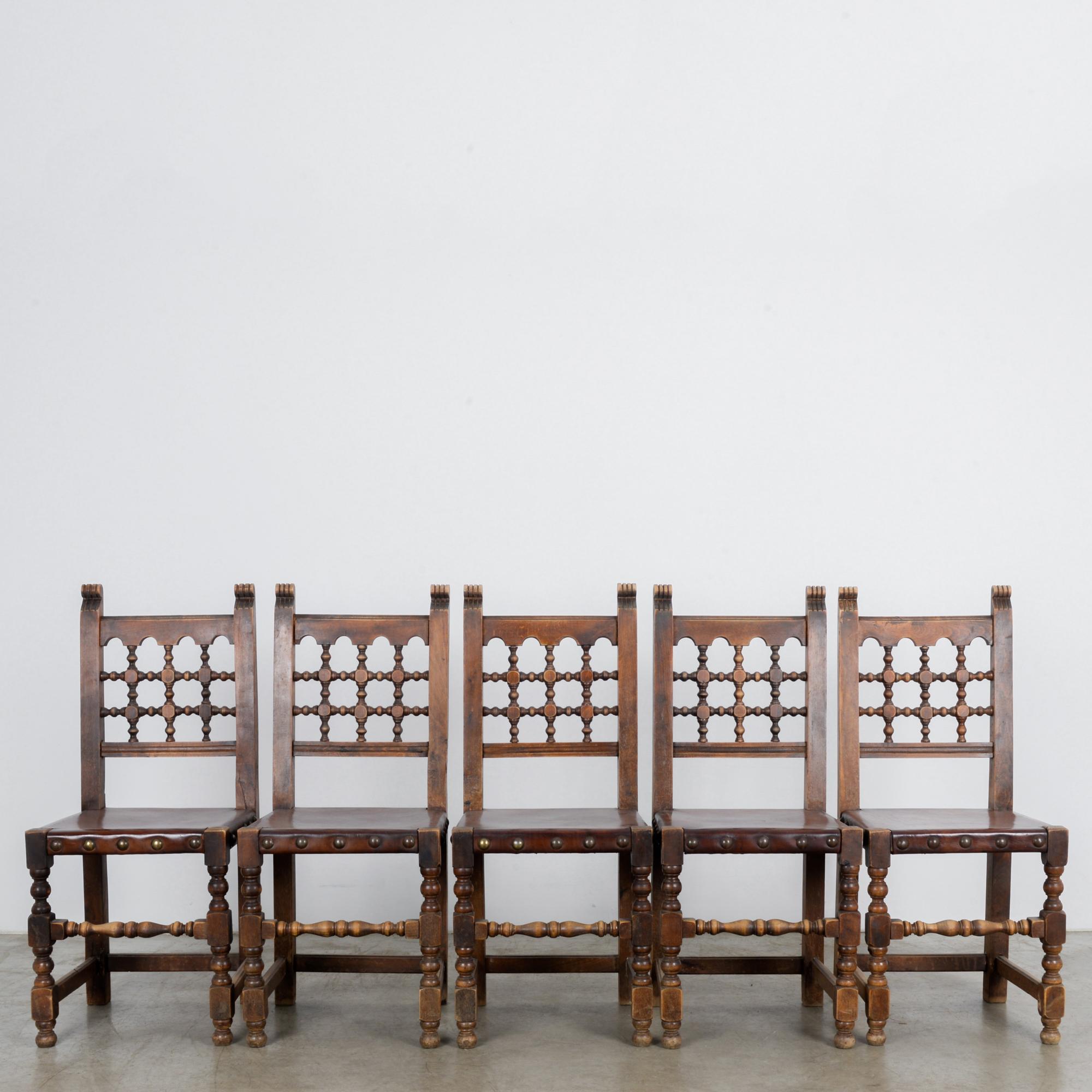 A set of five wooden dining chairs with leather seats from Belgium, circa 1960. Hearty, dark wood forms a straight outline for this contemporary take on Tudor style seating. Fine craftsmanship in the carving of the seat backs into quipu like grids