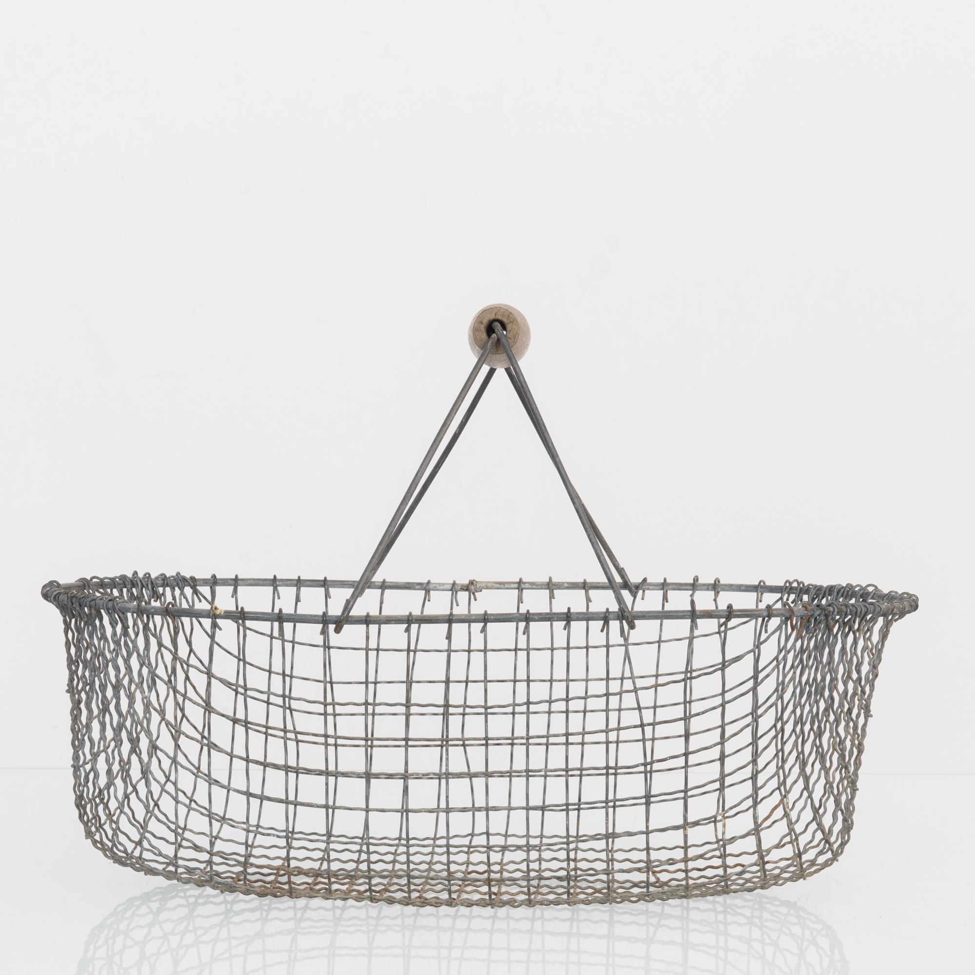 This basket was made in Belgium, circa 1960. Woven from thin metal wires, the basket displays a netted, organic structure with depth and a gentle flare. The pot shape is accentuated with heavy wire straps, clasped with a wooden handle.