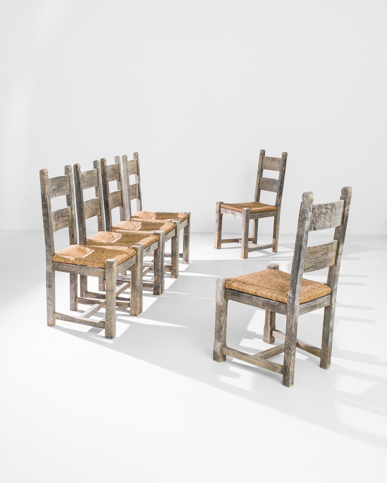 A set of six oak dining chairs from 1960s Belgium. The farmhouse shape is accentuated by the rustic finish of the wood, marked by a textural grain. A woven rattan seat brings out the amber undertones beneath the natural silver-grey color of the oak,