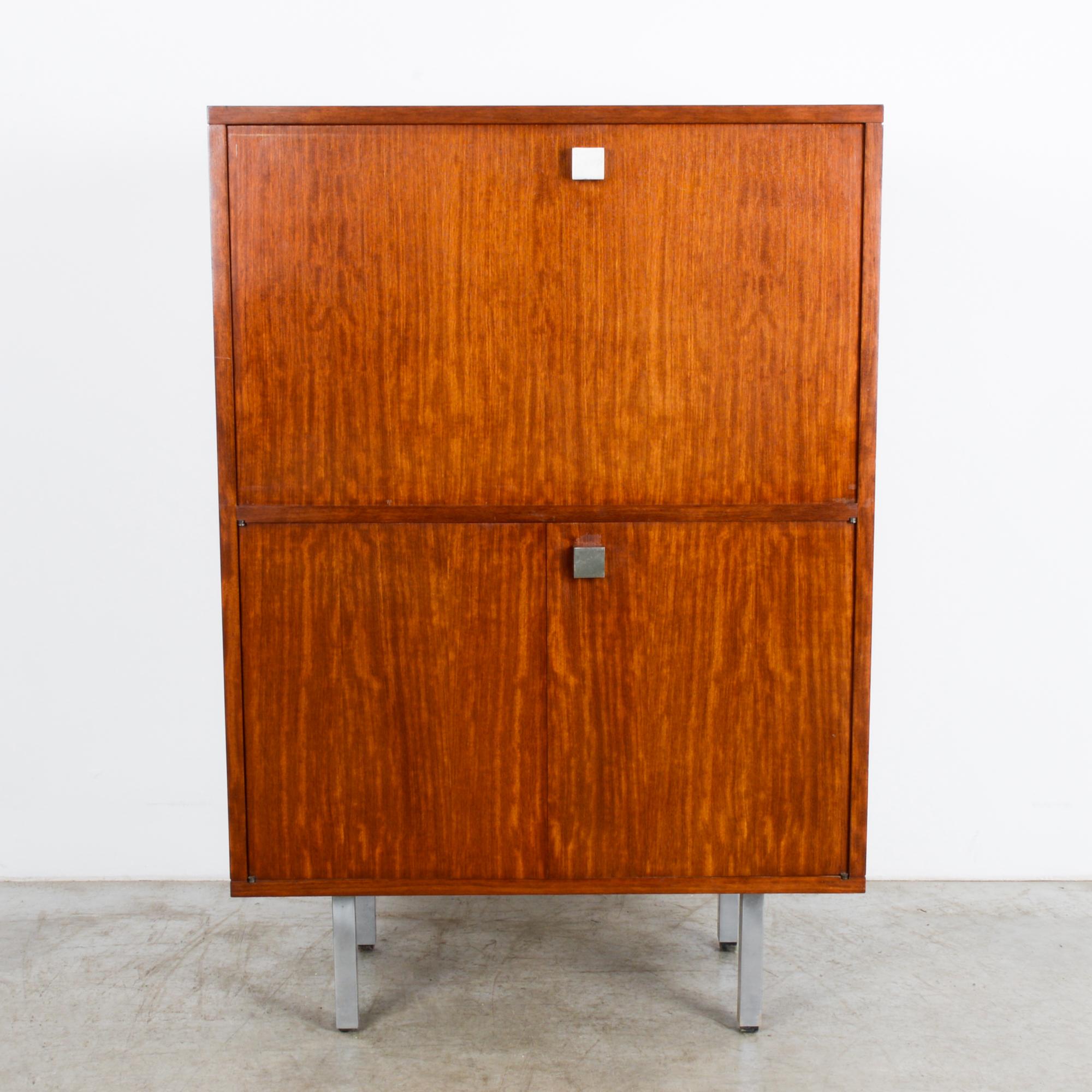 A teak cabinet from Belgium, circa 1960, from the studio of furniture designer Alfred Hendrickx. A cubic cabinet raised on simple silver legs, cleverly stores a concealed secretary desk. An upper door folds down to reveal a writing desk, complete
