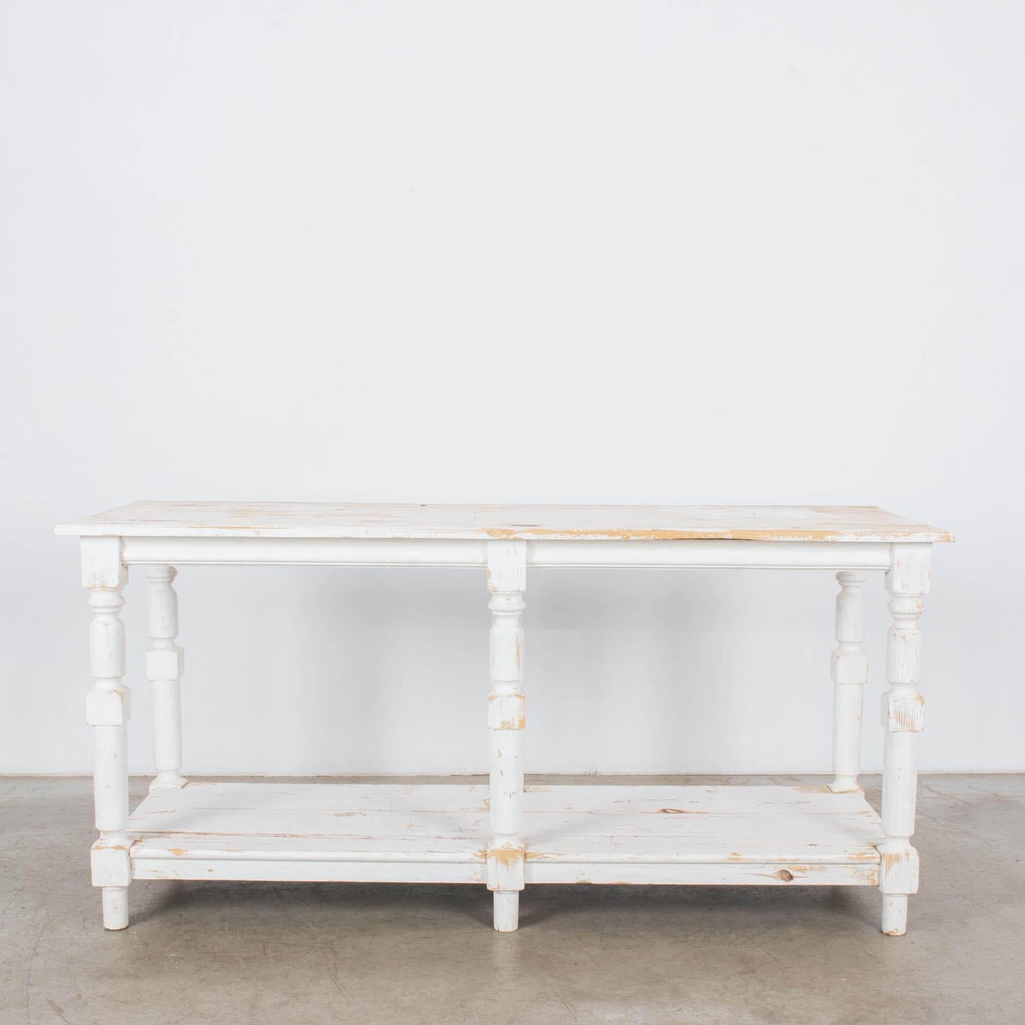 A wooden drapers table from Belgium, circa 1960. Six turned legs support a square tabletop; below, a lower platform serves as storage space. The table is painted a crisp, cheerful white. The paint has worn away along the lines of the grain to create