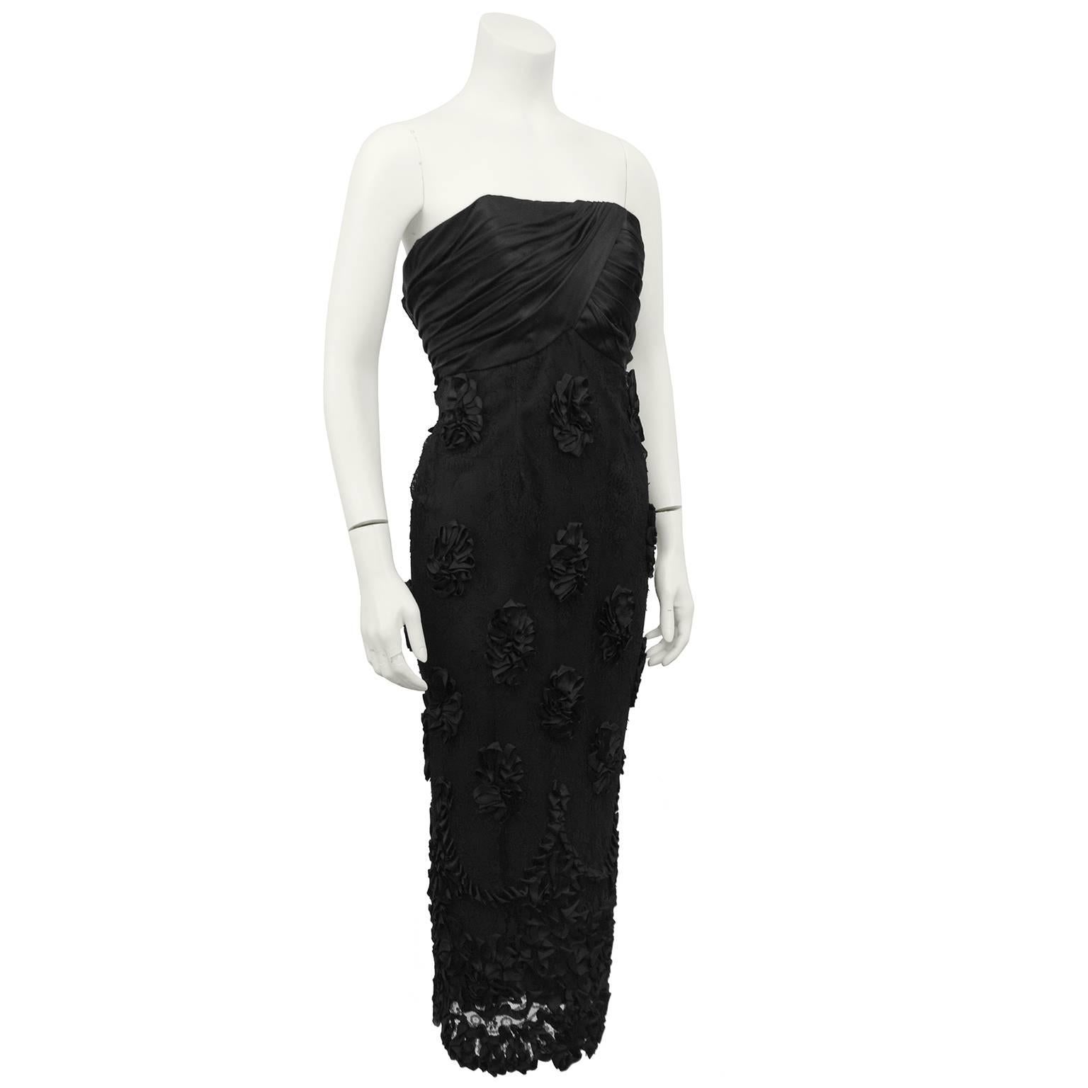 1960s black cocktail dress. Strapless and empire waist with a beautifully folded and ruched bodice. Straight, tea length skirt has a black lace overlay with large floral appliques made from black ribbons. More black ribbon applique details at bottom