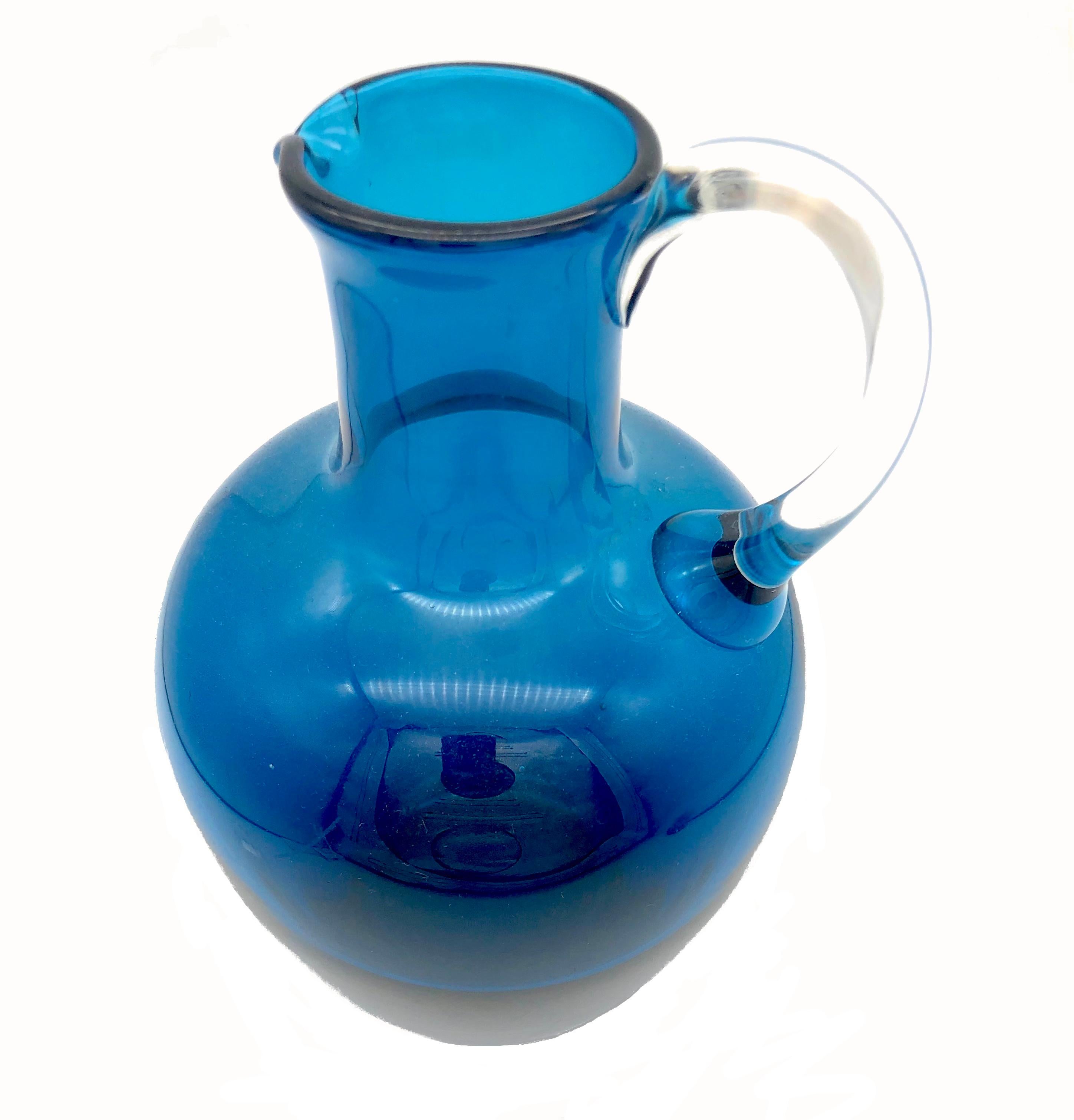 Dieser elegante blaue Glaskrug wurde um 1960 in Handarbeit hergestellt. Der elegante und schlichte Grundriss ist typisch für diese Zeit. Die blaue Farbe des Gefäßes beginnt unten dunkler und wird nach oben hin heller. Der transparente Henkel wirkt