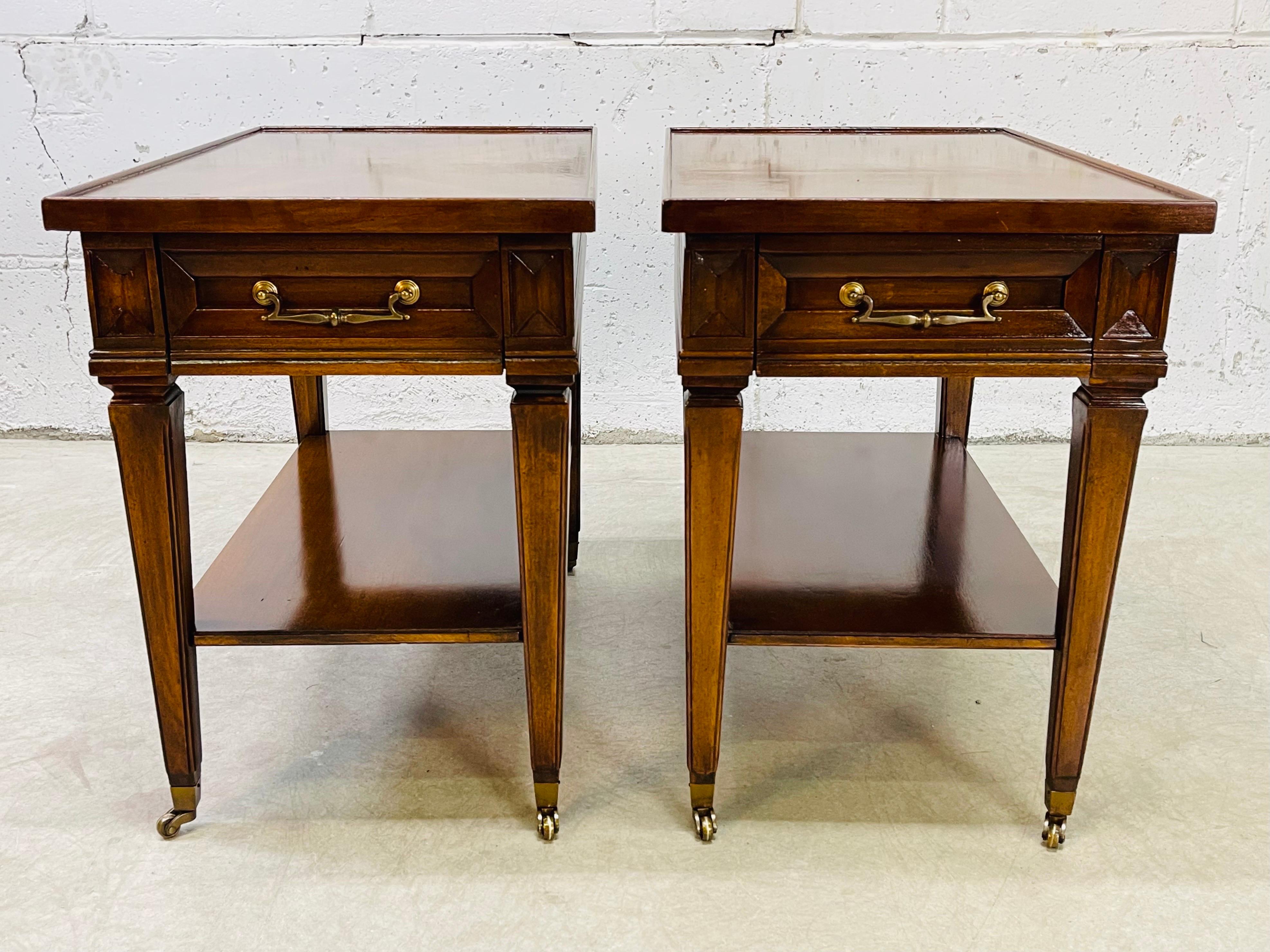 Vintage 1960s pair of mahogany wood side tables with a matchbook veneer top by Brandt Furniture Company. The tables have brass pulls and brass castors. The castors roll freely. Each table has a single drawer for storage and an additional shelf.