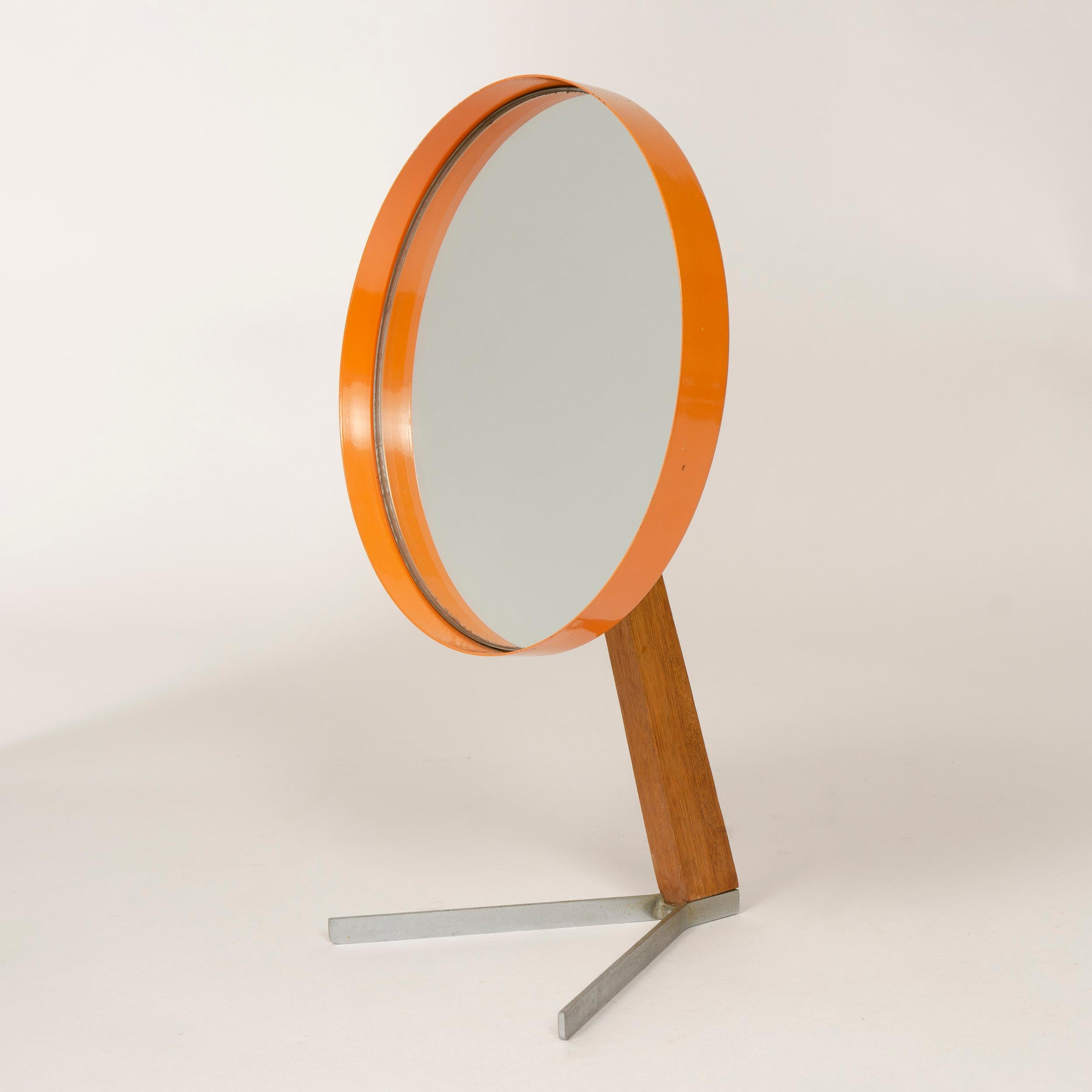 An adjustable orange enameled round table mirror rising from a wood pedestal and supported by two metal prong legs.