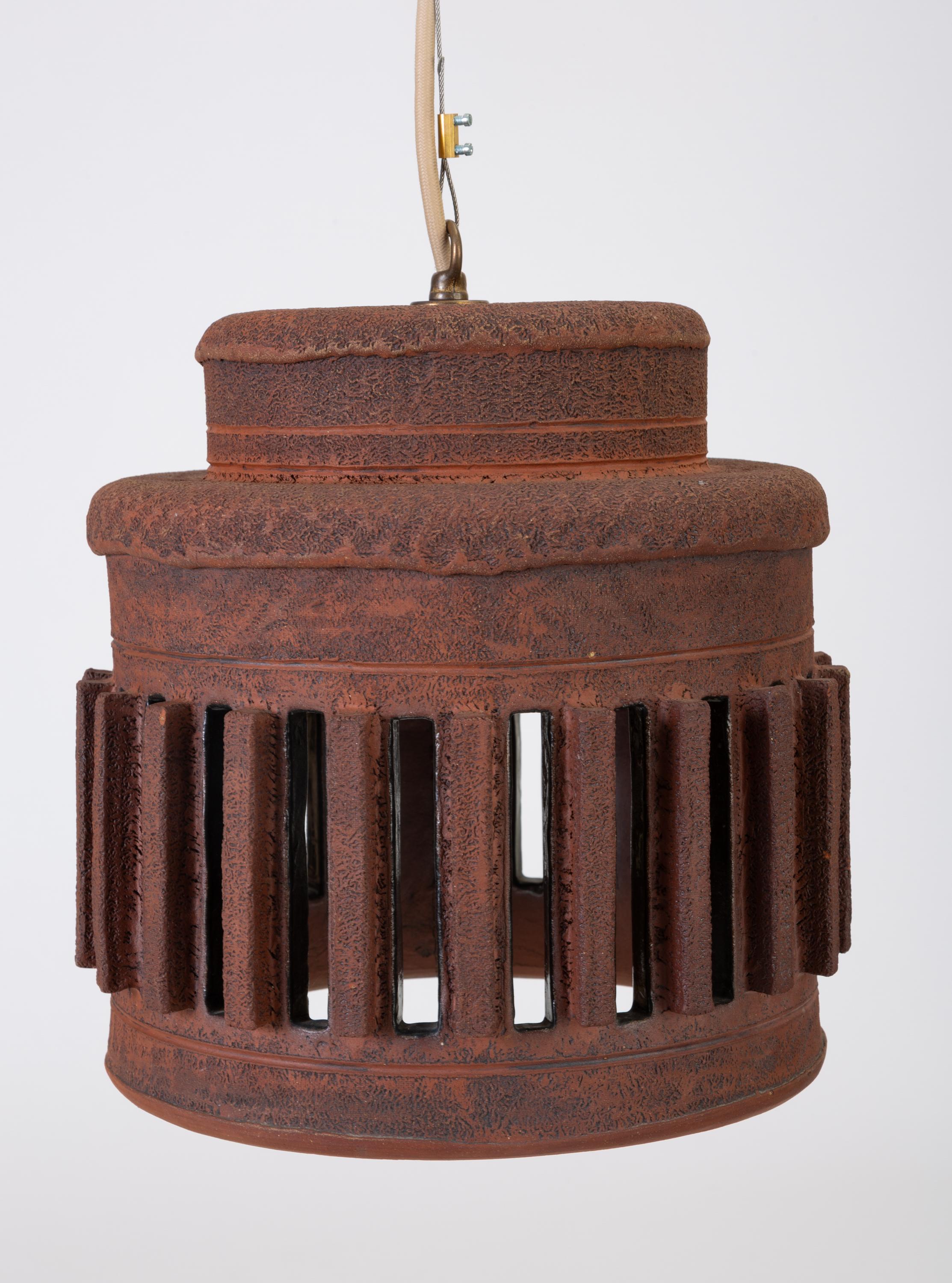 A unique stoneware pendant lamp with architectural, crenellated vents around the side. The lamp is made from a textured red-brown clay with handmade debossed and sgraffito detailing. This example has a rounded drum shape with raised vents. The light