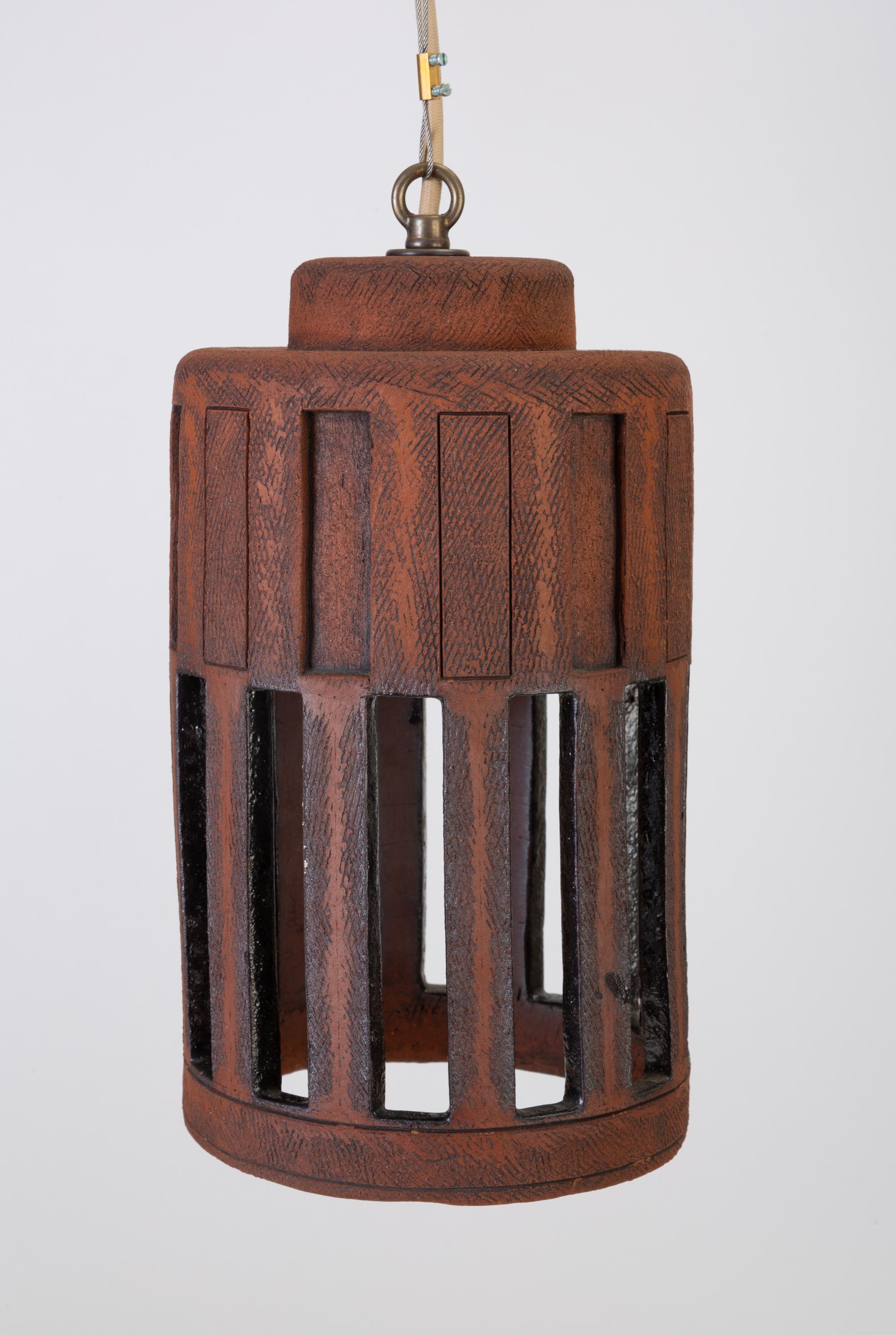 A unique stoneware pendant lamp with architectural, crenellated vents around the side. The lamp is made from a textured red-brown clay with handmade debossed and sgraffito detailing. This example has a long, narrow cylindrical shape with debossed