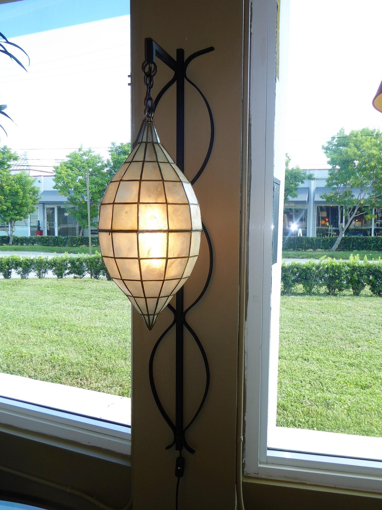 Unique and stunning, with a nod to Tony Duquette style, this wall sconce or light has a large Oblique Spheroid or teardrop form Capiz Shell pendant light hanging from a shaped wrought iron back, wall mounted. Completely restored, the wrought iron