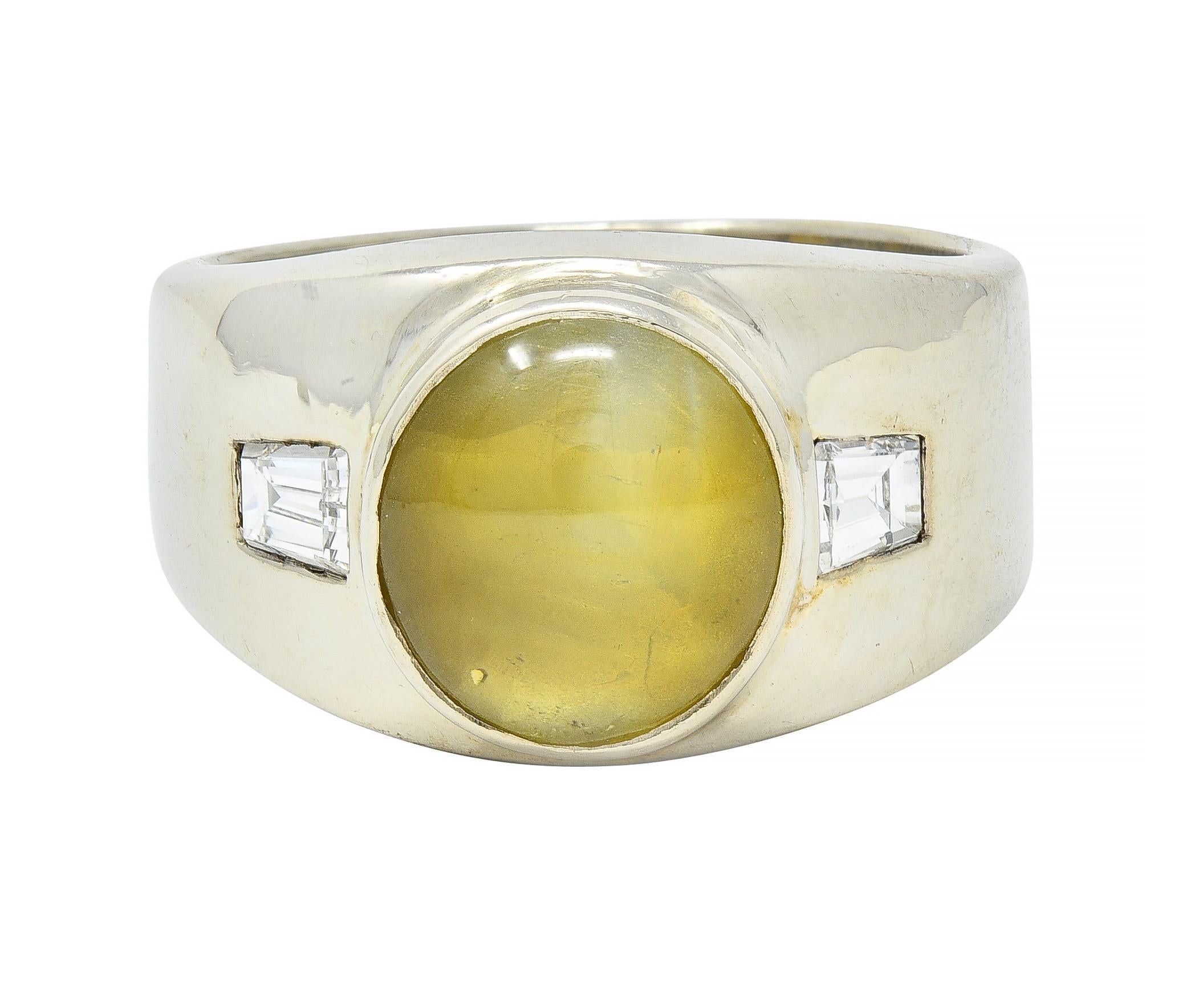 Centering an oval-shaped cat's eye chrysoberyl cabochon measuring 10.1 x 9.6 mm 
Yellowish green with linear chatoyancy and milk & honey effect - bezel set
Flanked by tapered baguette cut diamonds weighing approximately 0.36 carat total 
G color