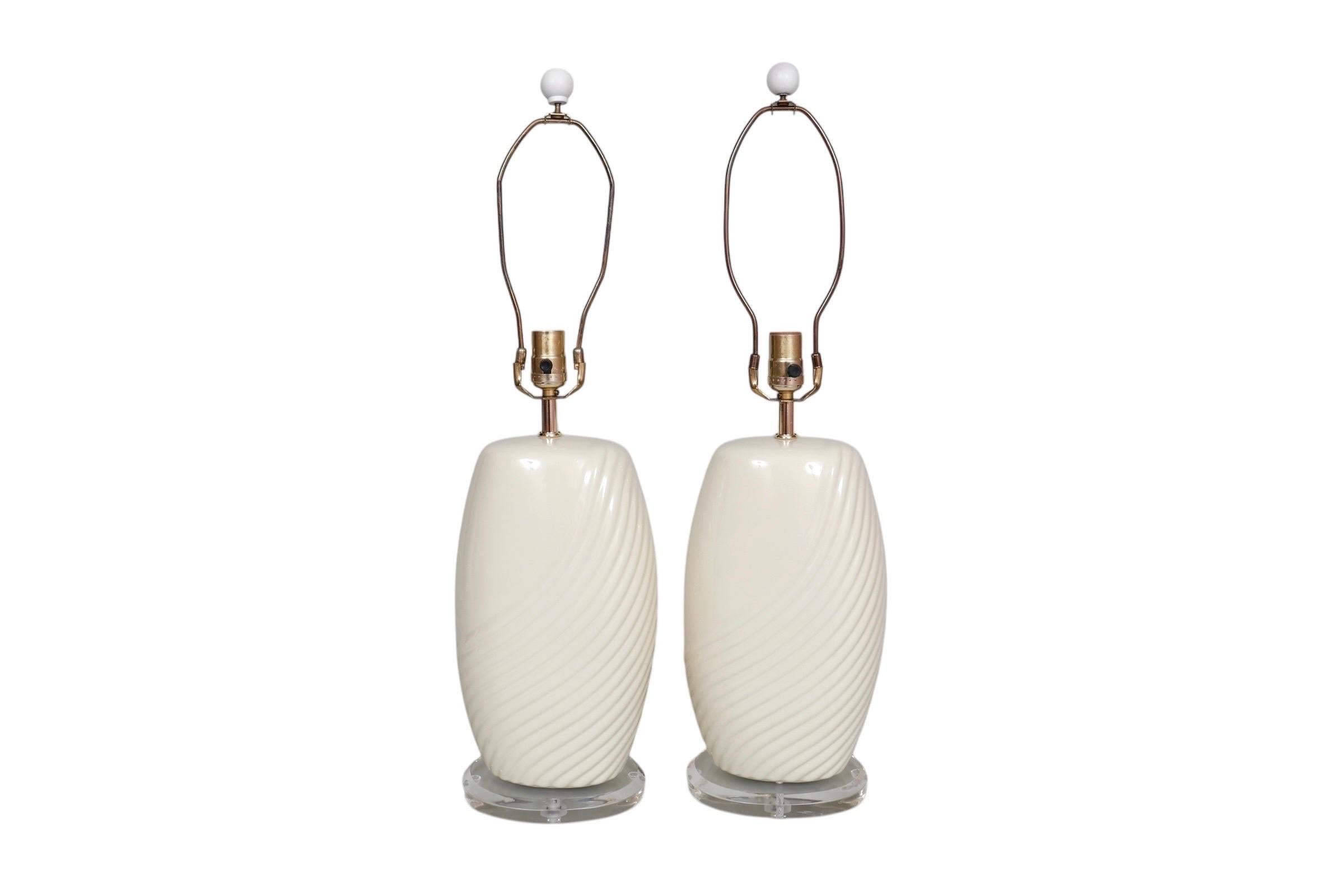 Paire de lampes de table en céramique blanc cassé, vers 1960. Les bases rondes biseautées en Lucite supportent des vases de forme ellipsoïdale, nervurés avec une vague sur une épaule. Les lignes légèrement incurvées se reflètent dans les plis