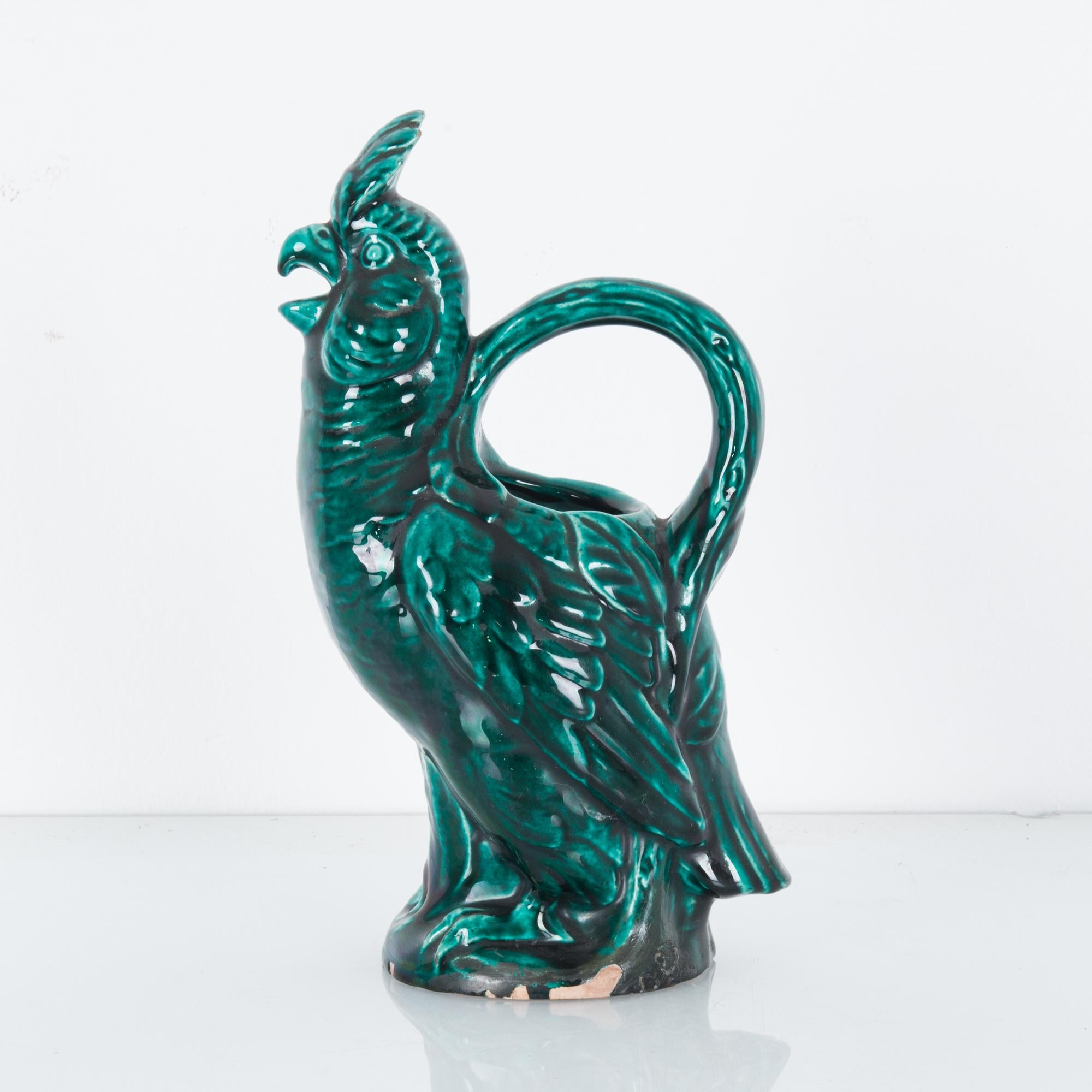This ceramic jug in the shape of a rooster was made in Italy, circa 1960. It has a carrying handle above its body and a beak spout. The rooster’s feathers and details are well rendered. With its glossy myrtle green glaze, the jug will be a charming