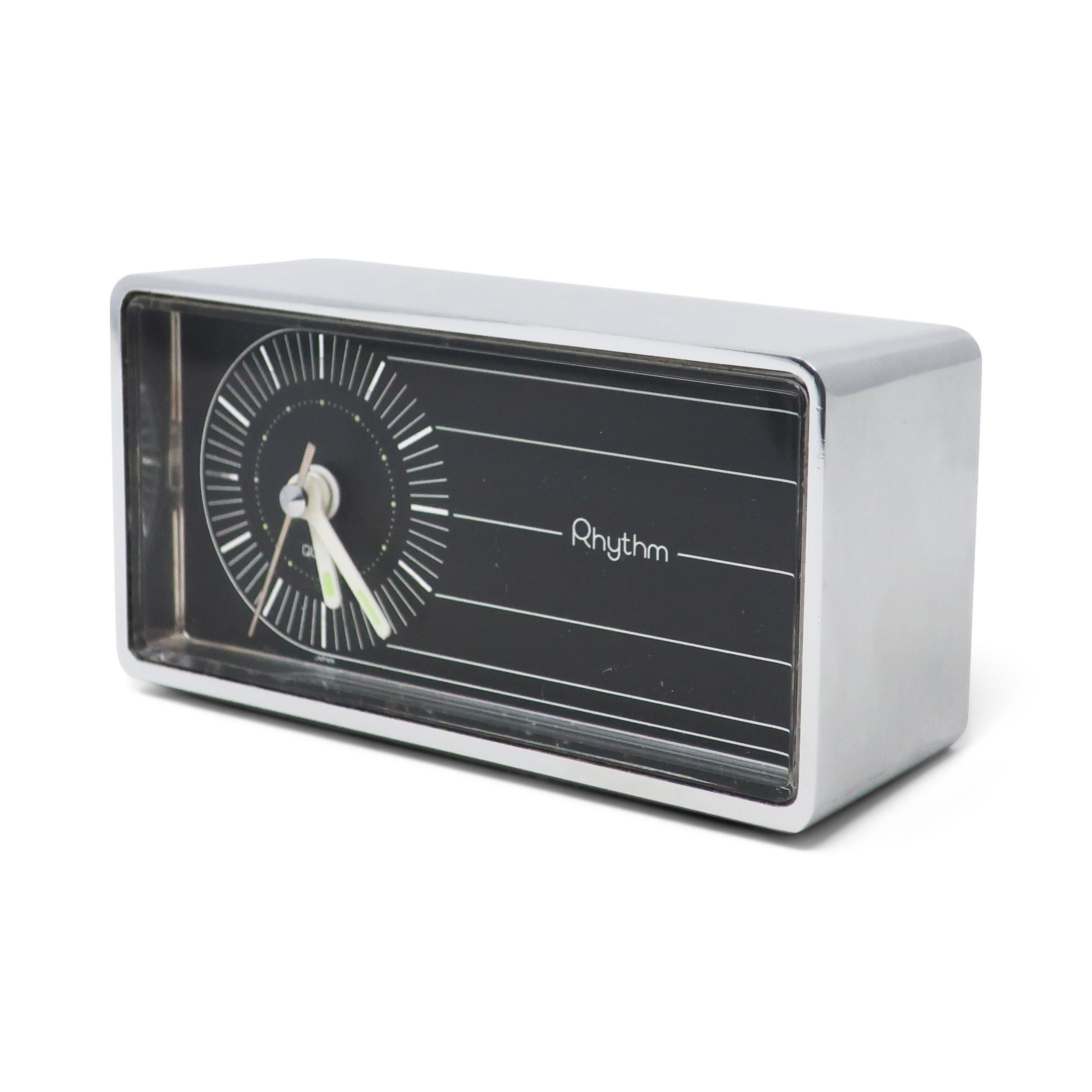 A perfectly Mid-Century Modern alarm clock by prolific Japanese clock manufacturer Rhythm. Rectangular chrome case with rounded corners, black face with white markings, white hands, and glow-in-the-dark paint on the hands. Takes a single AA