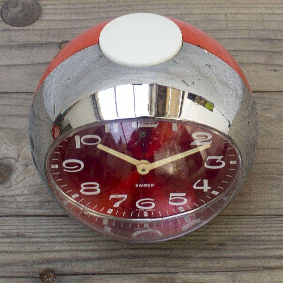 This 1960s Mid-Century Modern tabletop clock was manufactured by Kaiser in Germany. It features a silver chrome outside framing with a red iridescent clock face and large white numbering. The rounded shape adds to the midcentury design of the clock.