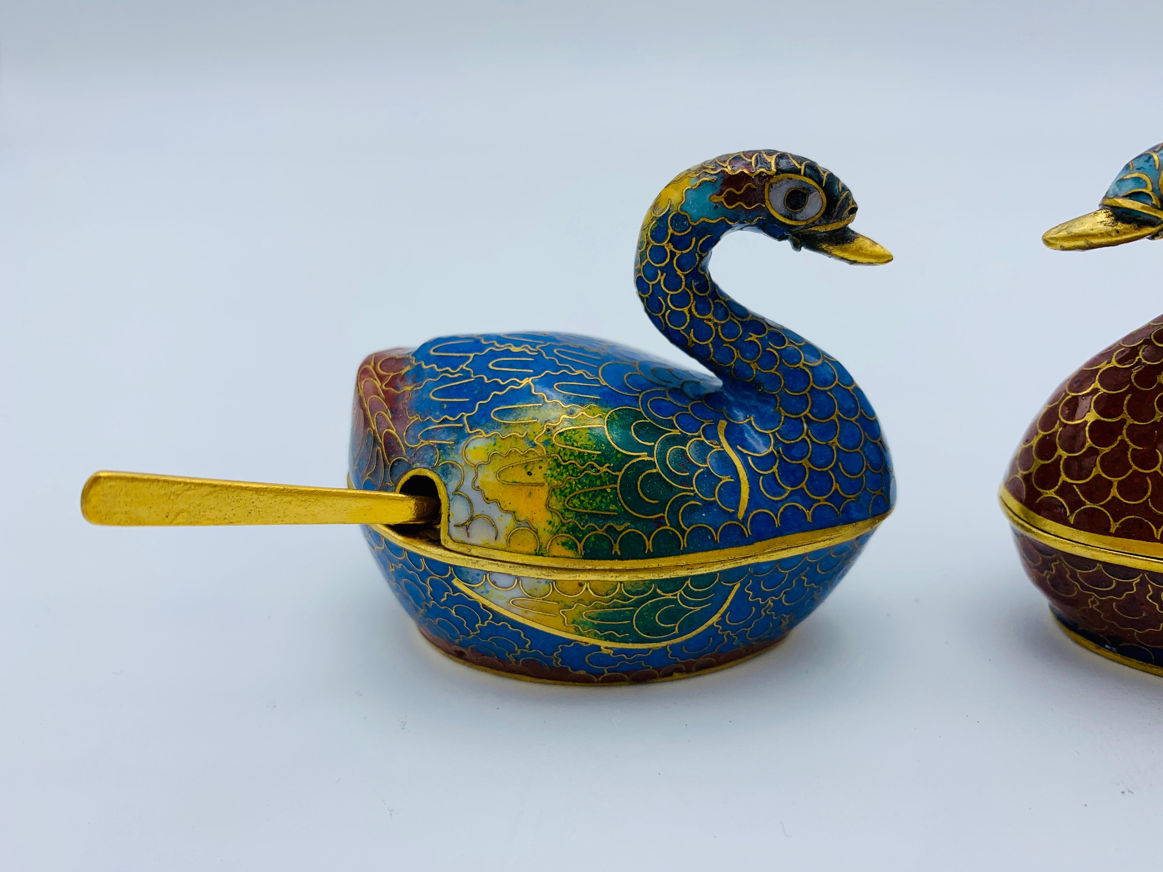 Listed is a beautiful and rare, pair of 1960s cloisonné sculptural swan salt cellars. The pair includes one blue/green and one red/black, each including small brass spoons.