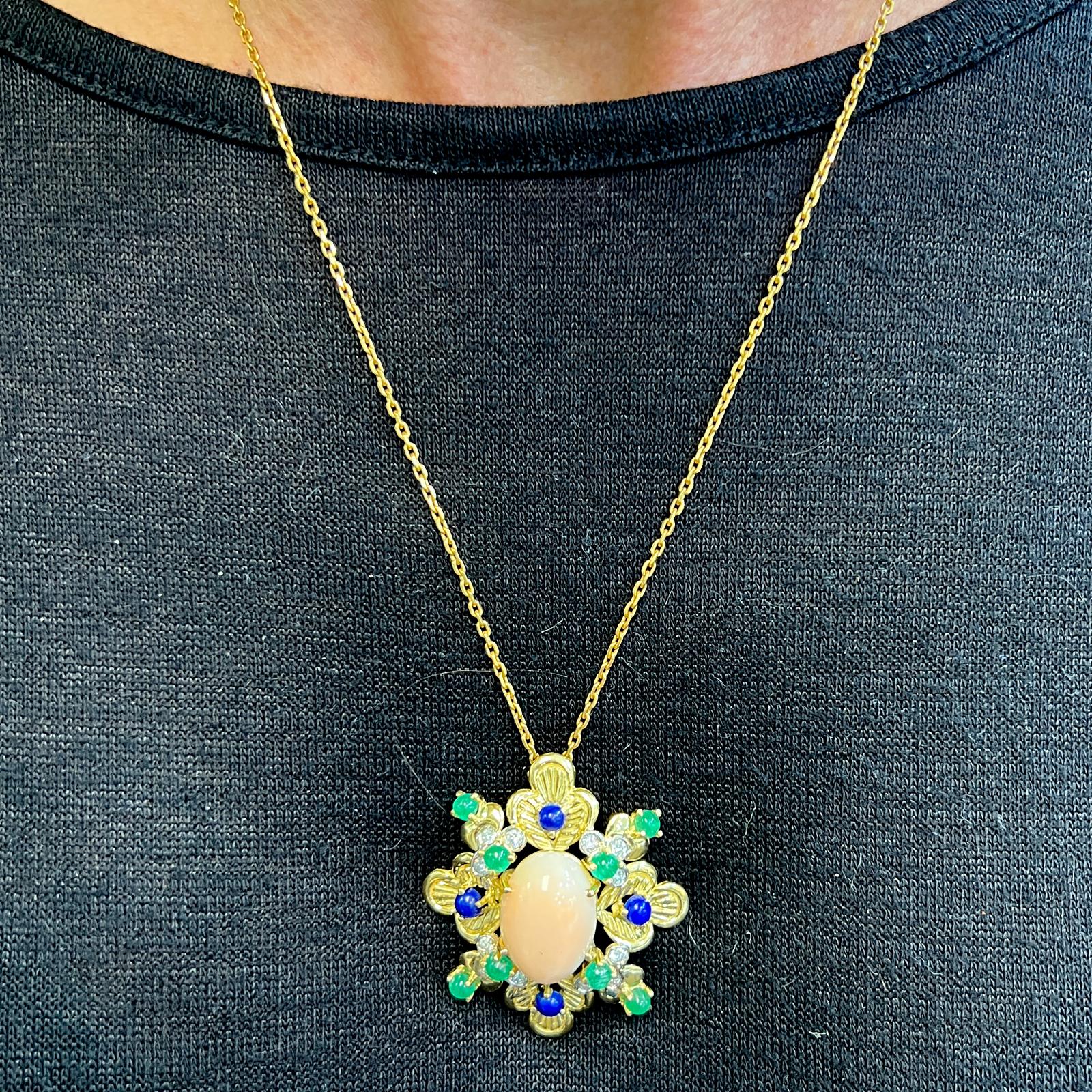 Beautiful coral, diamond, sapphire, and emerald pendant hancrafted in 18 karat yellow gold. The pendant features an oval cabochon coral gemstone surrounded by  4 sapphires,  8 emeralds, and  12 round brilliant diamonds. The diamonds weigh