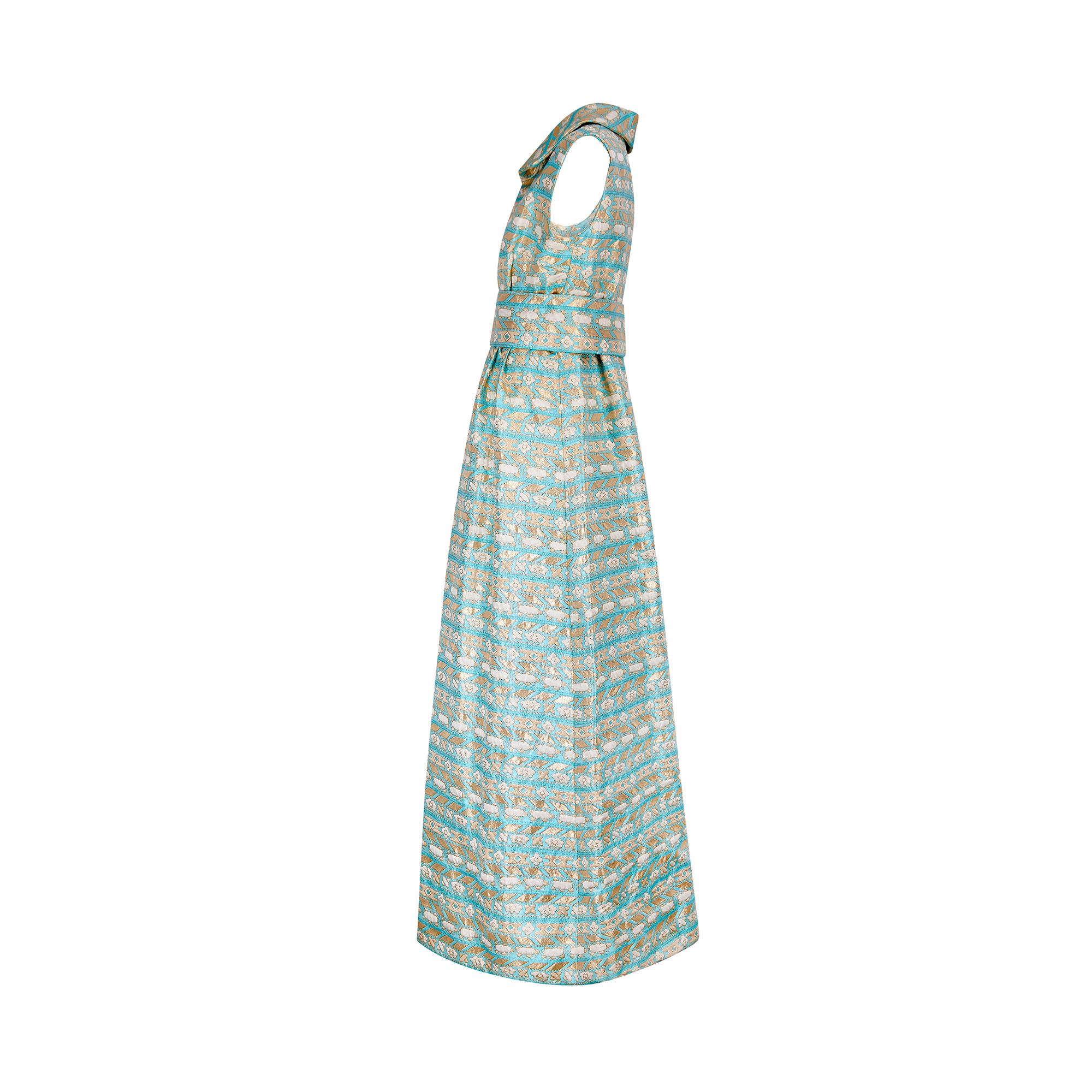 This striking gown was couture crafted in the late 1960s to early 1970s. The fabric is really the star of the show; thick aqua jacquard is textured with geometric, floral and cloud-shaped embroidery in glittering metallic gold. This dress comes