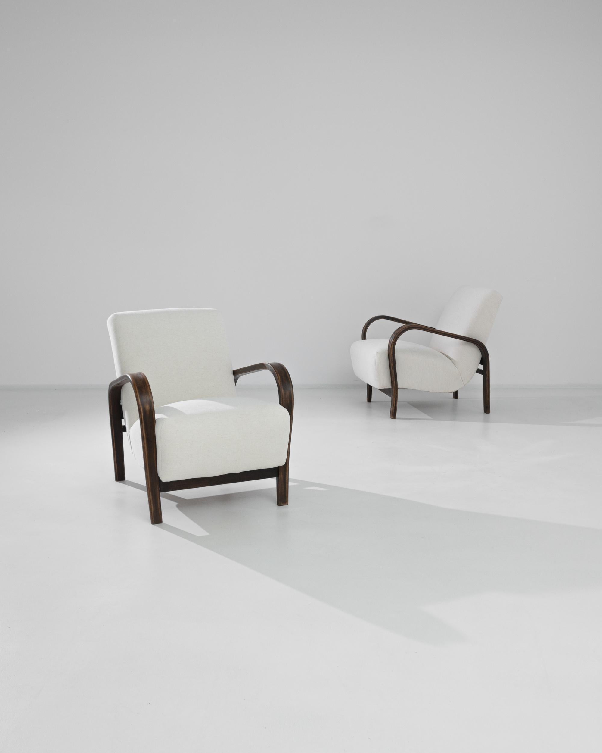 A pair of upholstered armchairs designed by Karel Kozelka and Antonin Kropácek, produced in Czechia circa 1960. Plush white curves of the cushions contrast beautifully with the dark brown of the bentwood armrests. This stylish pair is the best of