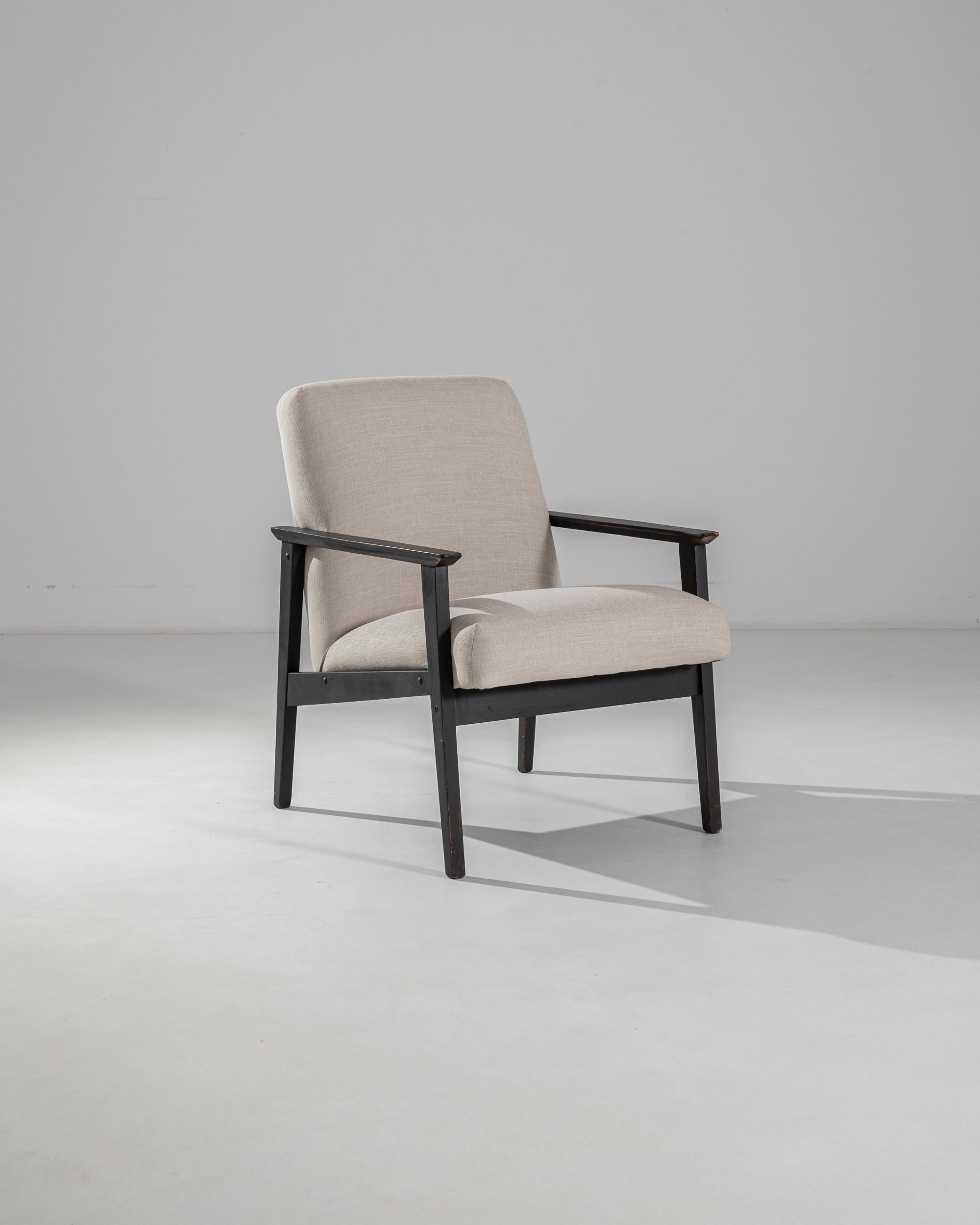 This upholstered armchair was produced in Czechia, circa 1960. Upholstered with a beige linen blend, the slight recline and cushioned seat make for a comfortable posture. Contrasting with the angular dark coffee frame, the plush seat creates a