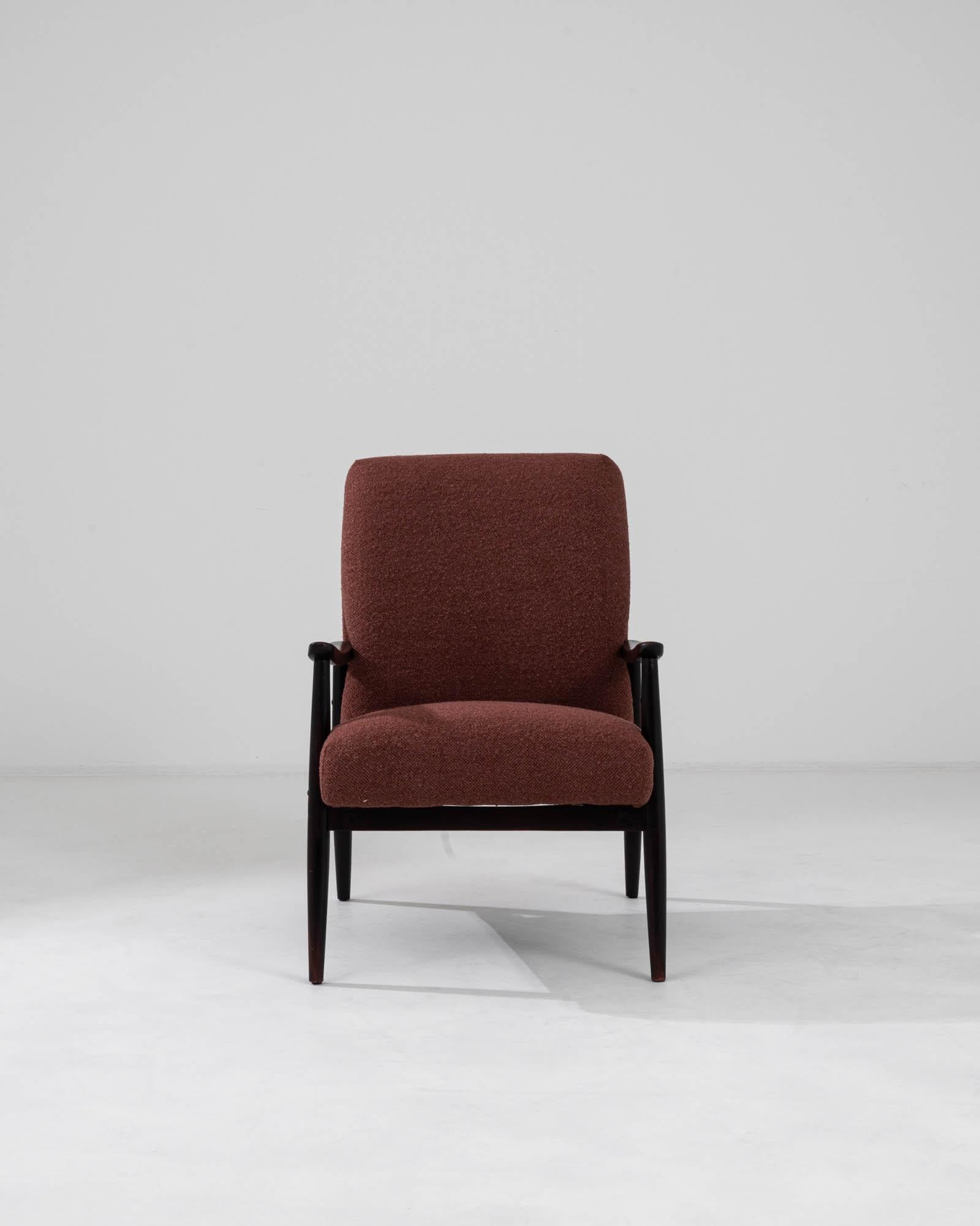 This upholstered armchair was produced in Czechia, circa 1960. Upholstered with a garnet fabric, the slight recline and cushioned seat make for a comfortable posture. Contrasting with the angular dark frame, the plush seat creates a subdued