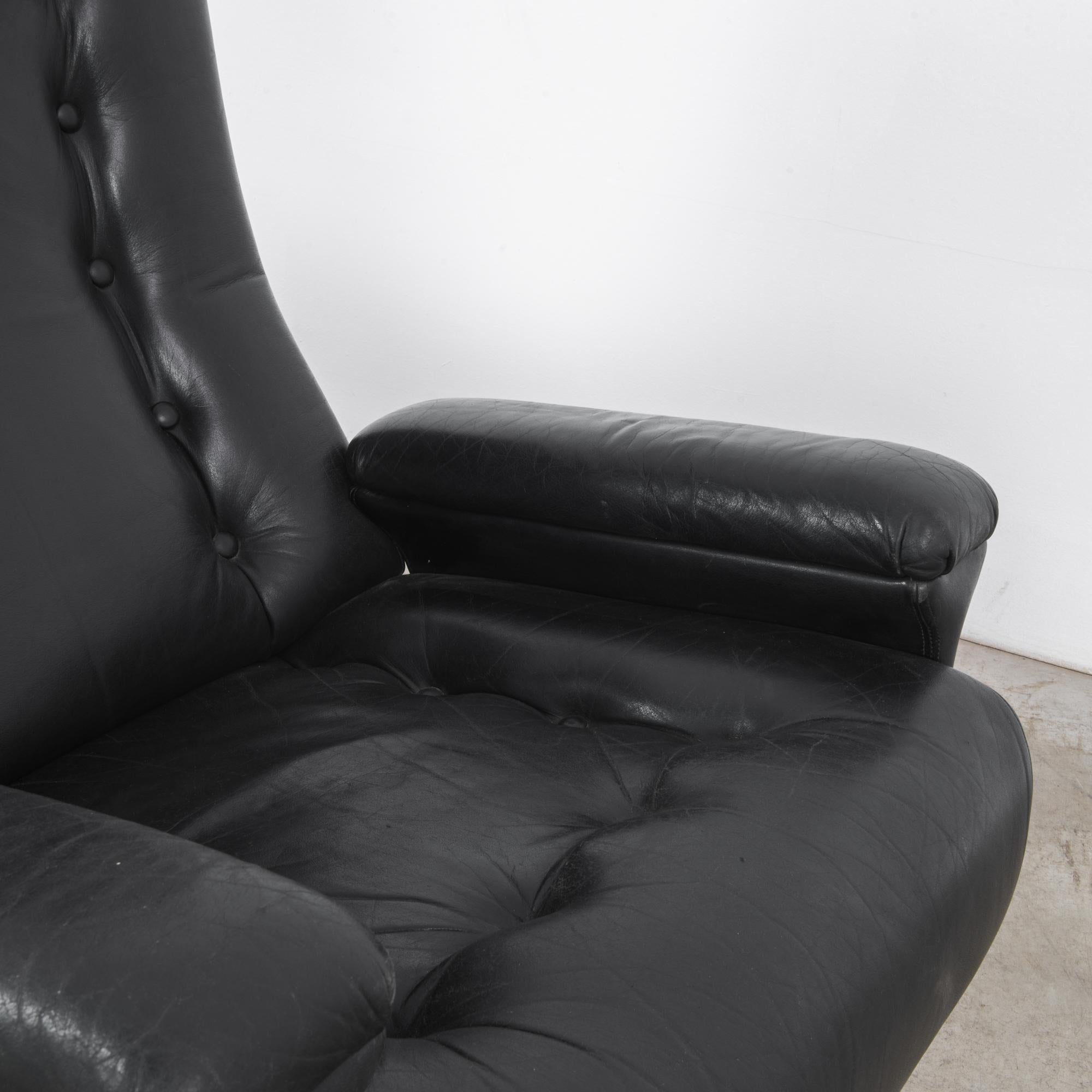 A pair of black leather armchairs from Denmark, produced circa 1960. A plush pair of midcentury loungers standing on five feet in rich black leather framed by wood panel side supports. Their assertive profiles make them perfect as comfortable accent