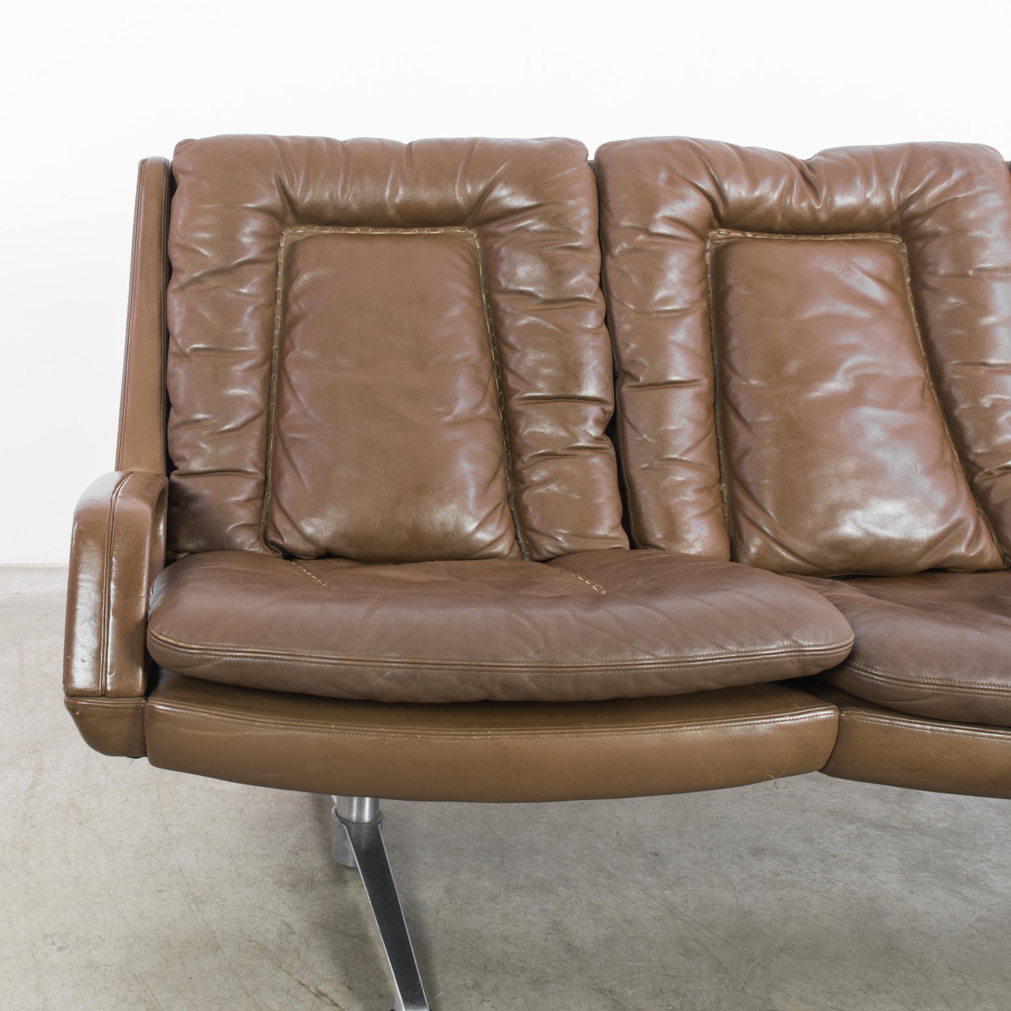 A leather sofa from Denmark, circa 1960. The design has a modernist lounge aesthetic; three chocolate brown leather seats are united into a wide bench, elevated upon minimalist silver feet. The comfortable curve of the seats and the plush leather