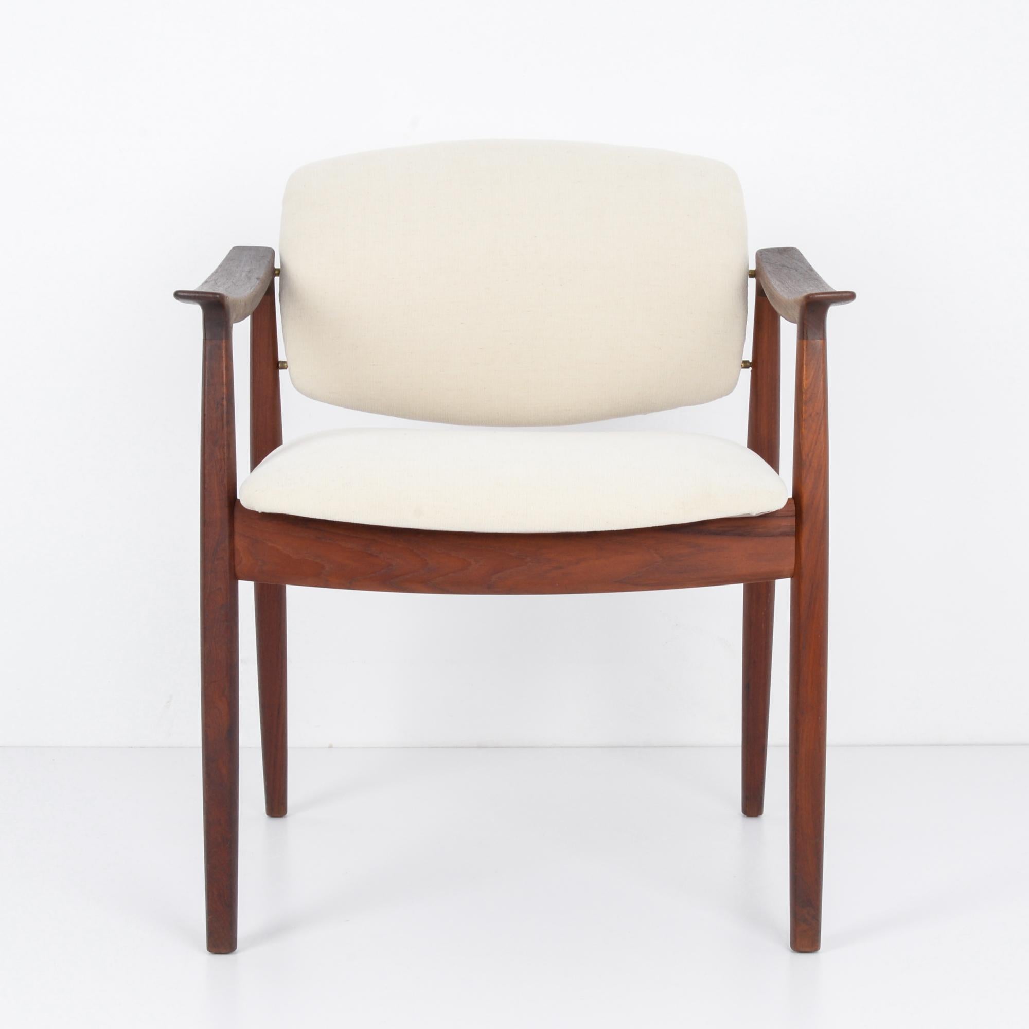 A wooden armchair from Denmark, circa 1960. Dark, mocha-colored wood with a soft polish, and a seat and backrest upholstered in a natural white. The frame is low-set, the back legs are set at an elegant slant, and the slope of the armrests invites a