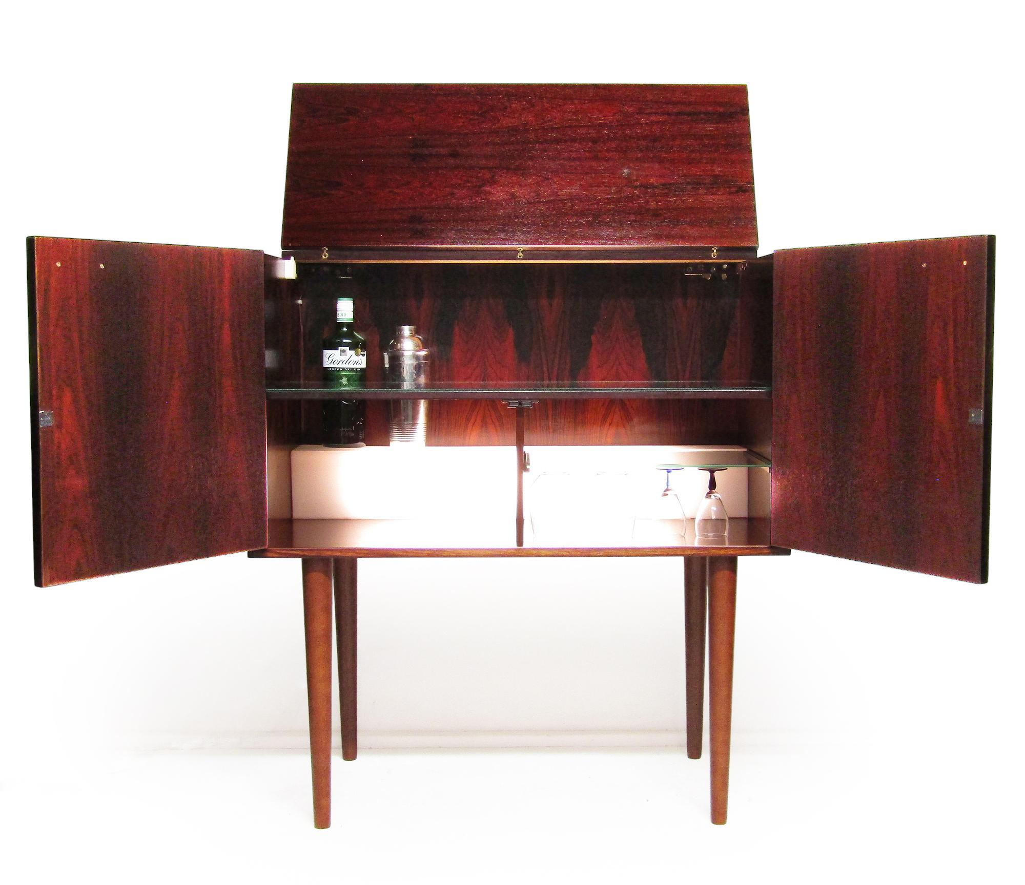Una elegante vitrina de cócteles de los años 60 en palisandro de Río, de Kai Kristiansen para FM Møbler.

Dispone de iluminación interior, estantes de cristal, una superficie de preparación y un estante iluminado para botellas.

Una luz alargada, de