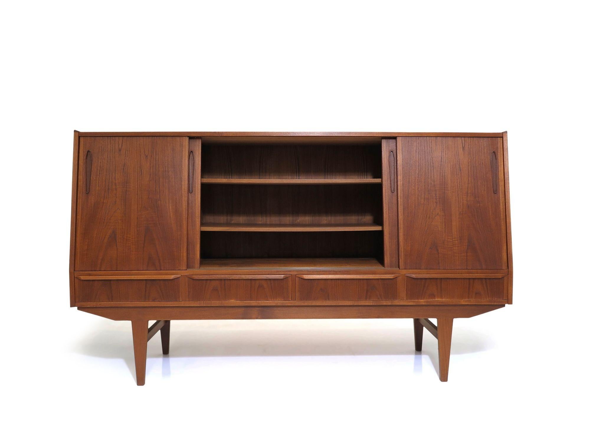 Scandinavian tall credenza with angled front crafted of old-growth teak with book-matched sliding doors revealing an interior with adjustable shelves and mirrored bar on left, above series of four drawers, raised on solid teak tapered