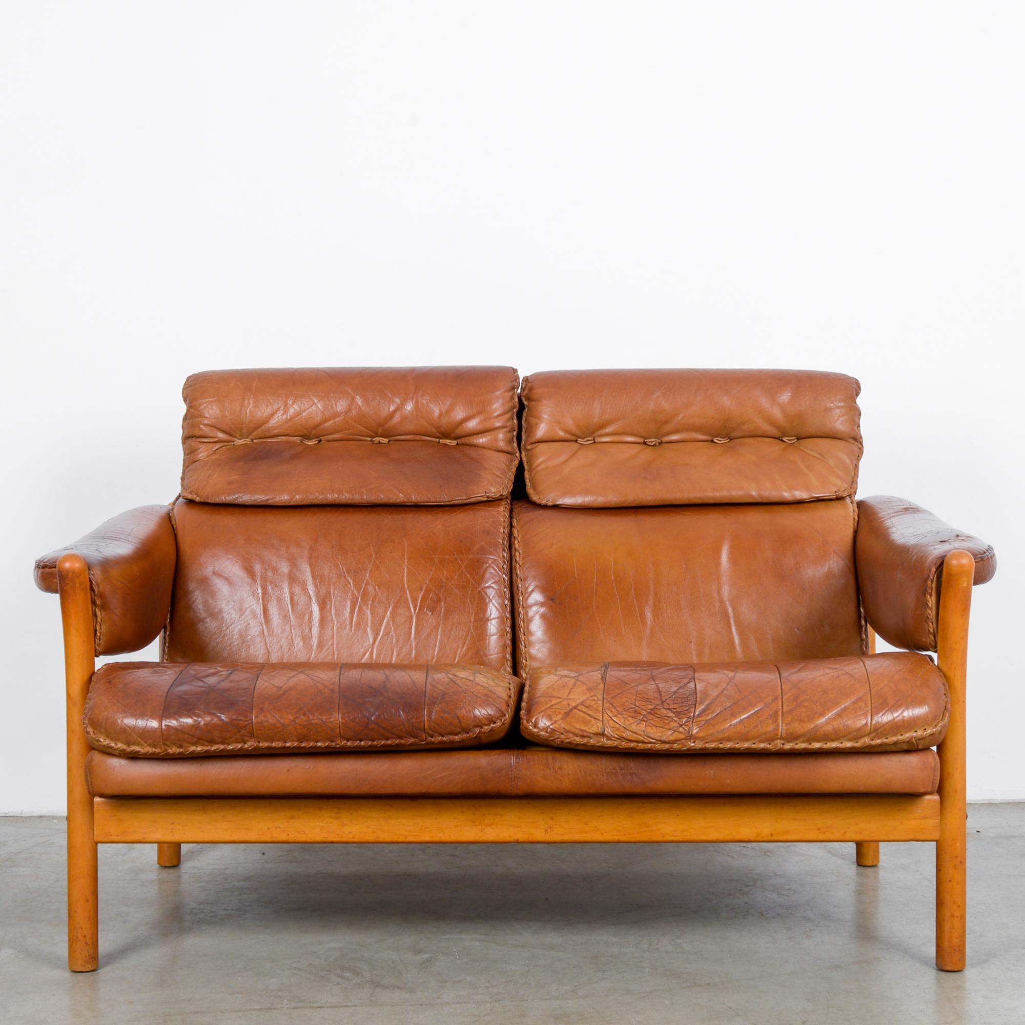 This two-seat leather sofa was made in Denmark, circa 1960. A wooden frame, gently curved, supports thick leather cushions-- while the golden yellow hue of the wood complements the warm brown tones of the leather. Stitched with leather strips,