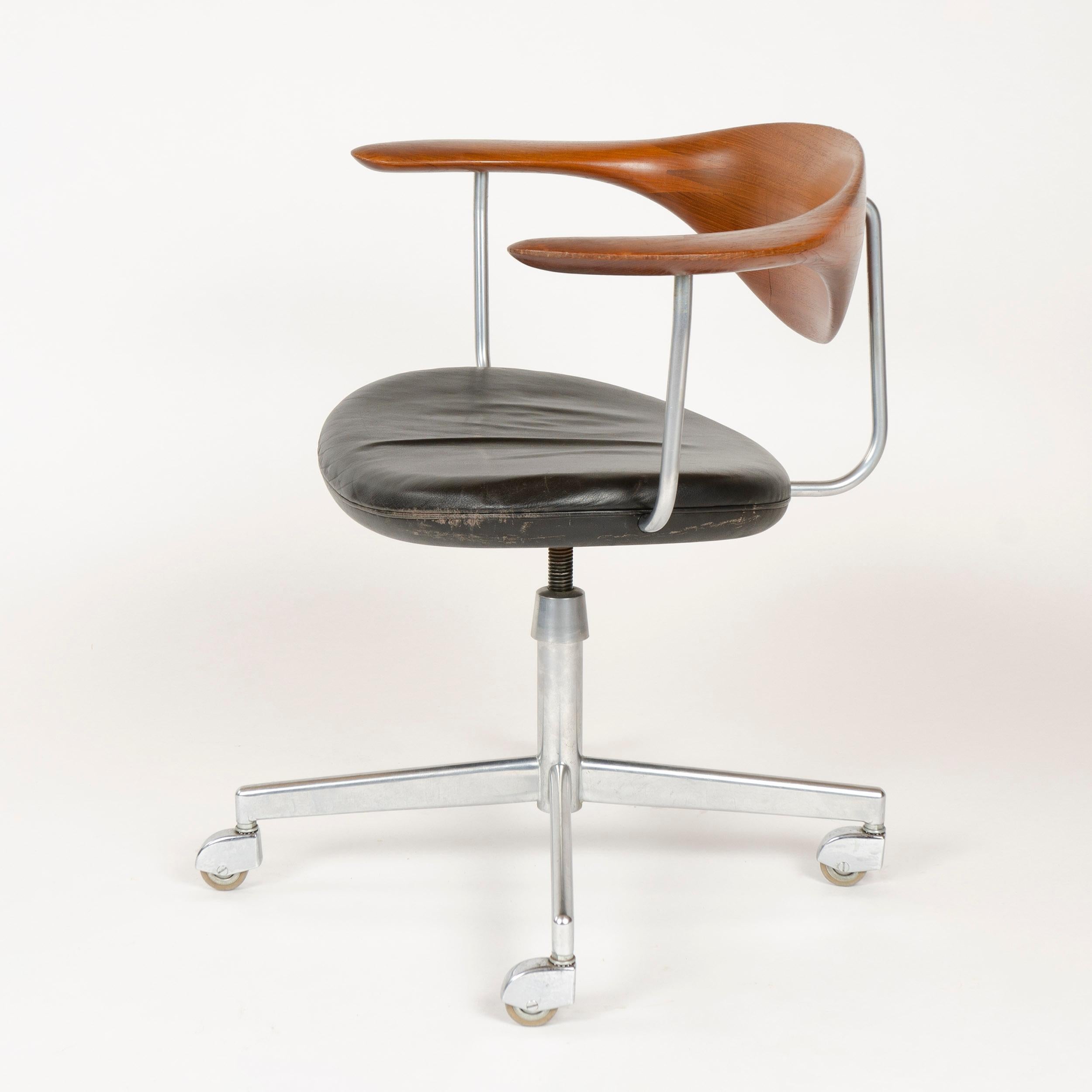 A superb swiveling desk chair having a hand-sculpted floating teak backrest and black leather seat, on a chrome base with hooded casters.